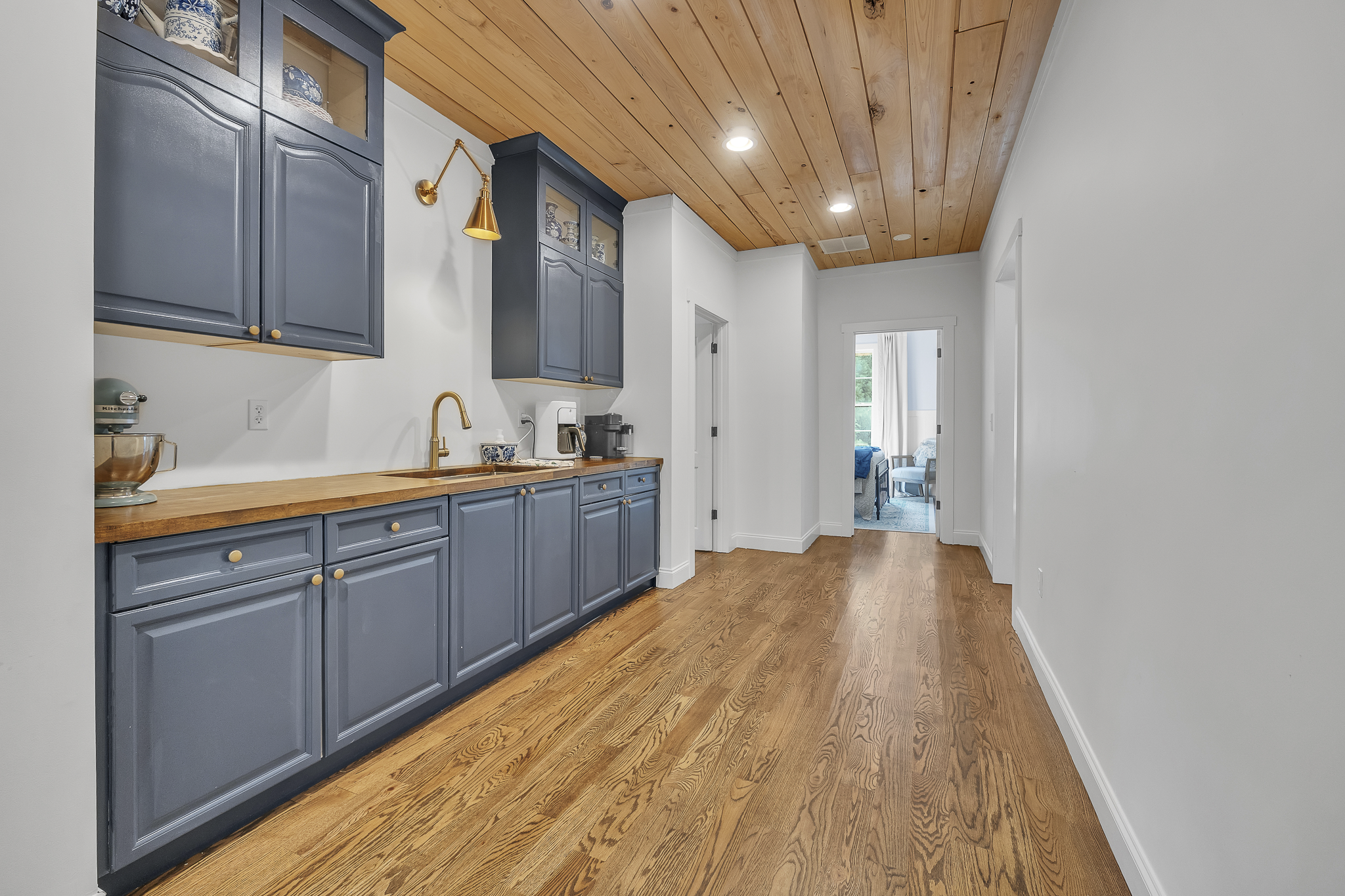 A hallway with wooden flooring, white walls, and a wooden ceiling. Blue kitchen cabinets with gold knobs along one wall, with a countertop holding a coffee machine, a bowl, and a mixer. There are three ceiling lights and entry doors at the end of the