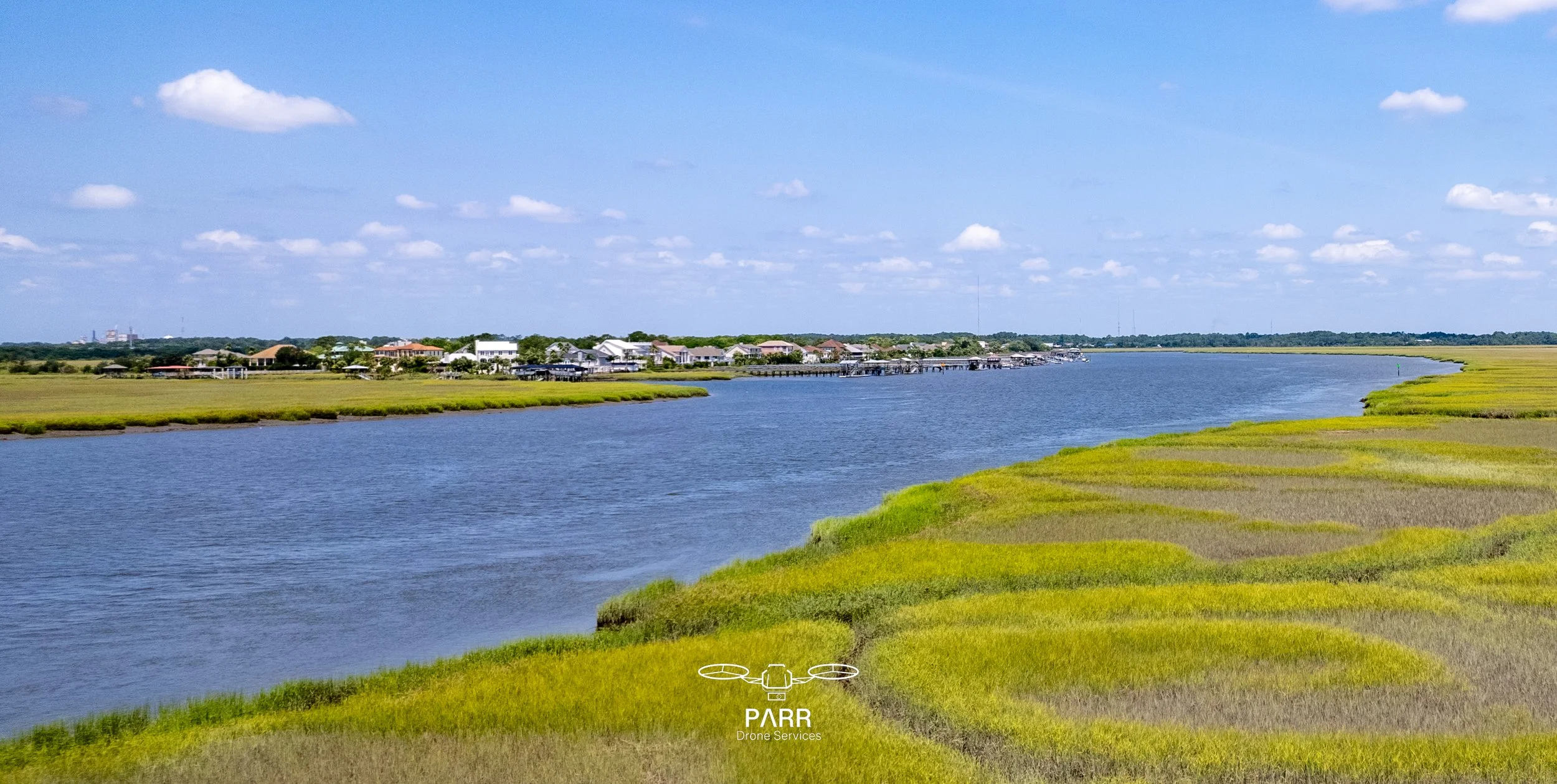 A landscape featuring a river with green marshes on both sides, a residential area with houses and docks in the distance, and a blue sky with scattered clouds.