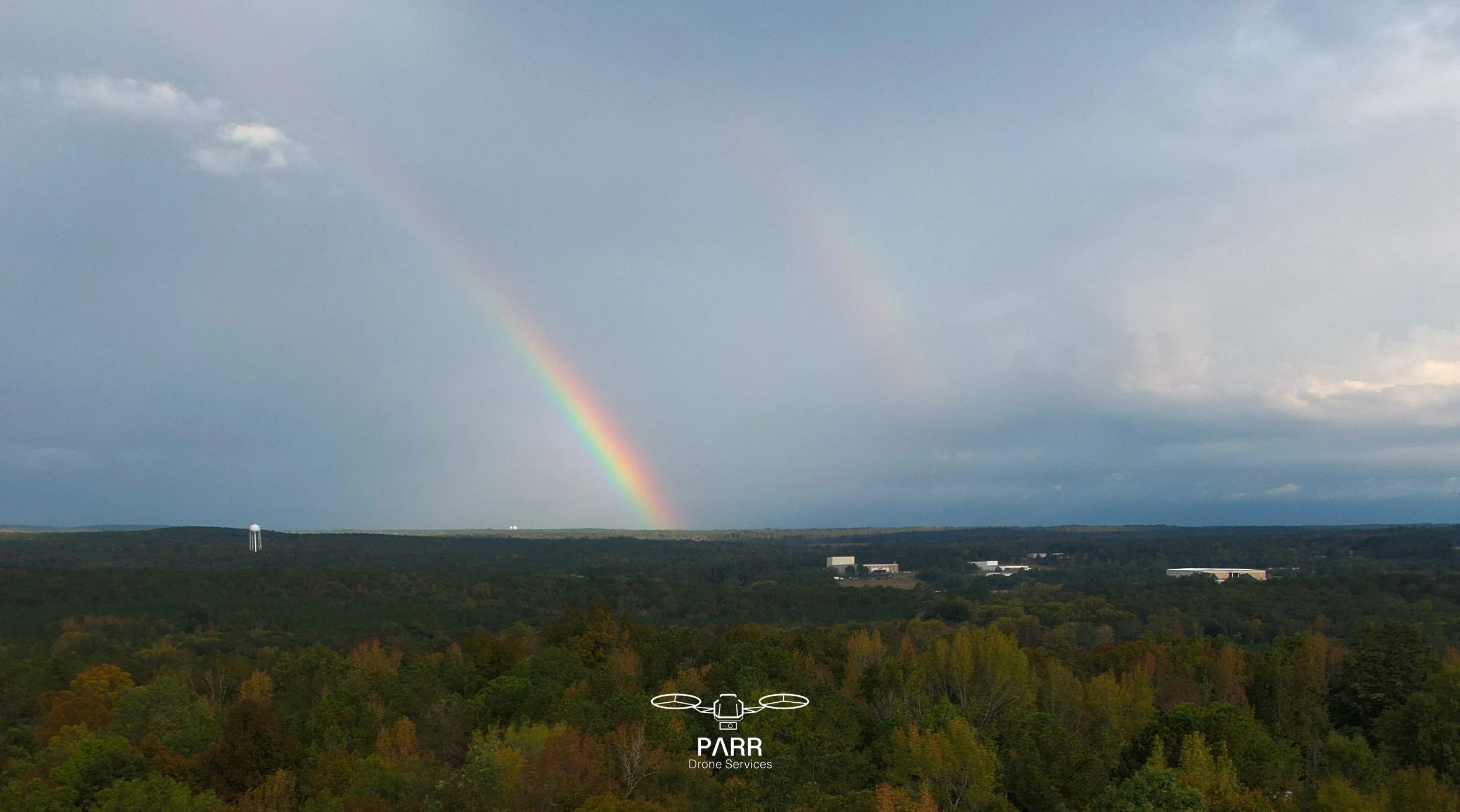 View of a colorful rainbow over a forested landscape with cloudy sky, buildings in distance, and a water tower, with a logo for PARR Drone Services at the bottom.