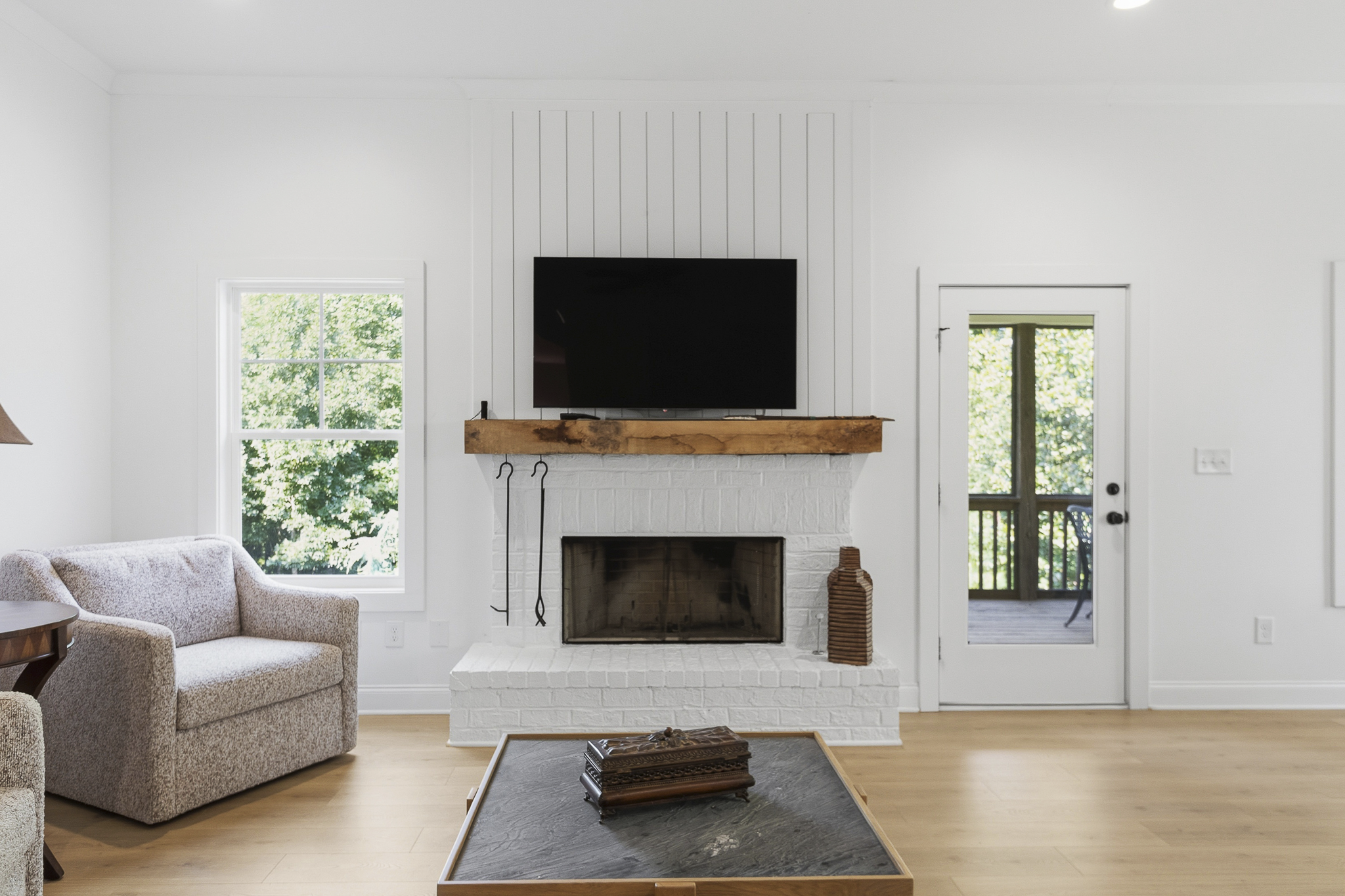 Living room with white walls, a brick fireplace with a wooden mantle, a flat-screen TV mounted above, a window on the left showing green trees outside, a glass door on the right leading to a balcony or porch, a beige upholstered sofa on the left, and