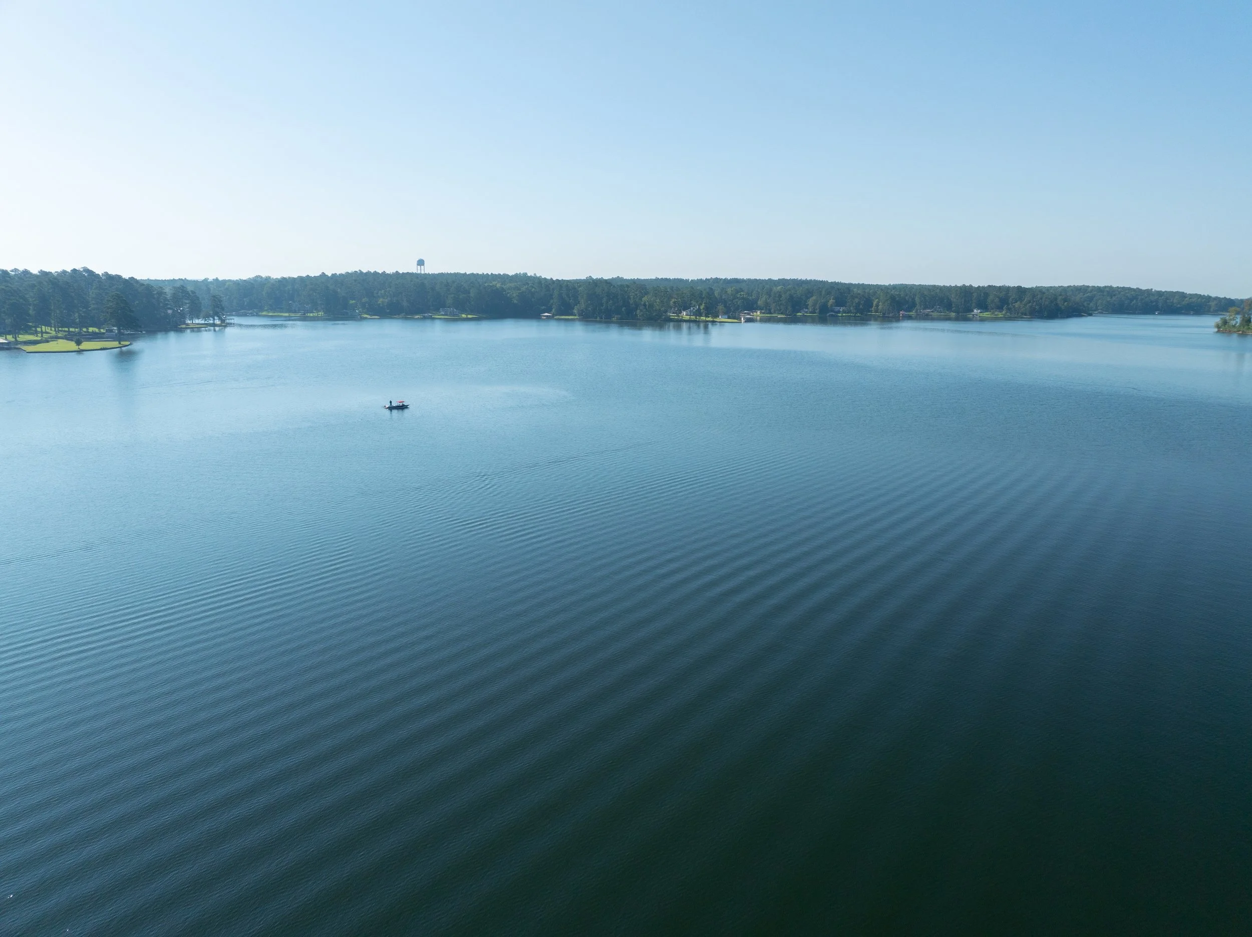 A large body of water with ripples, bordered by green treetops and a clear blue sky, with a small boat on the water.