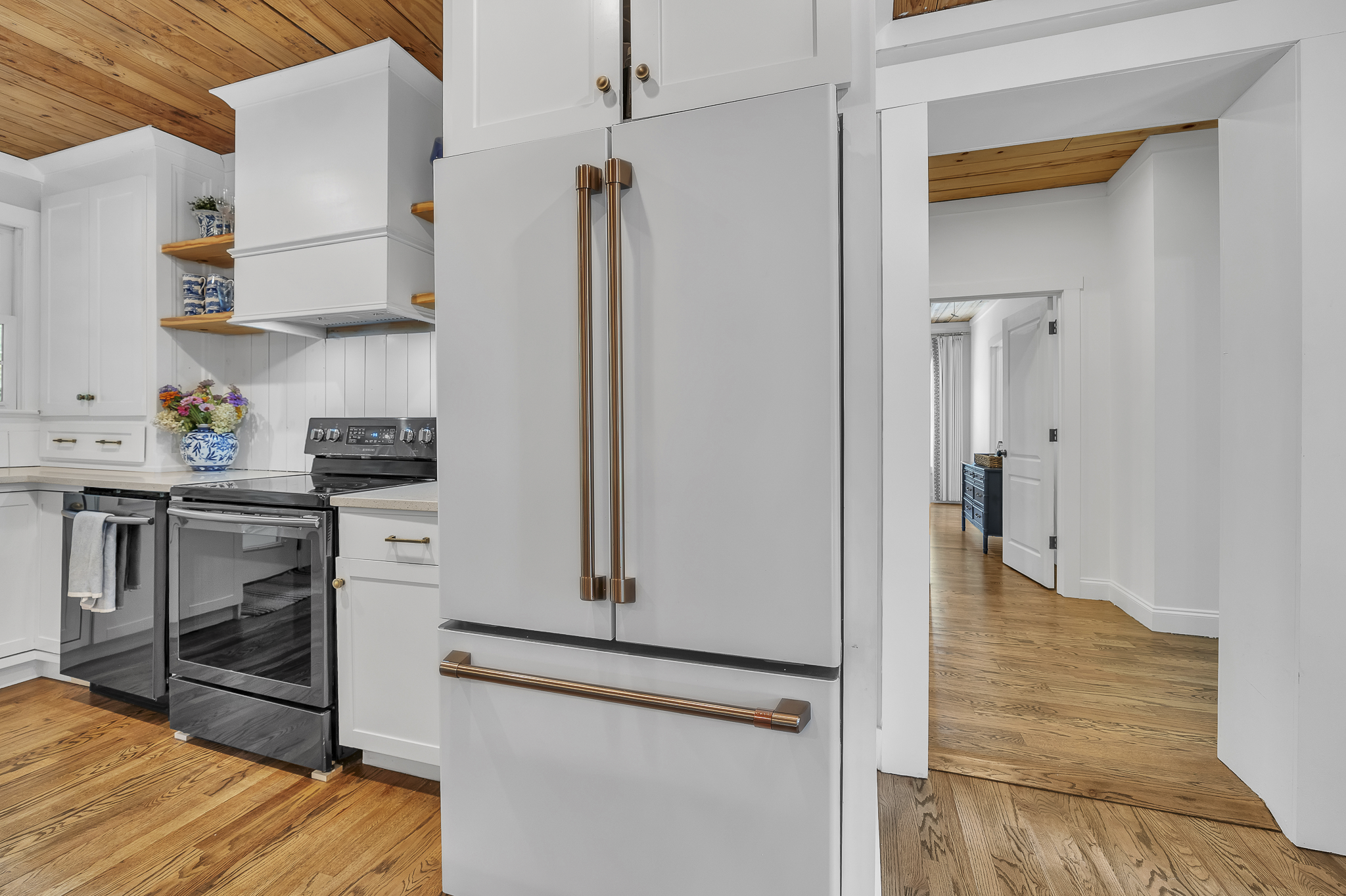 A kitchen with white cabinets, a black stove, and a large white refrigerator with copper handles. There are wooden open shelves with blue patterned dishes, and a blue and white flower vase on the counter. The ceiling is wooden, and the floor is hardw