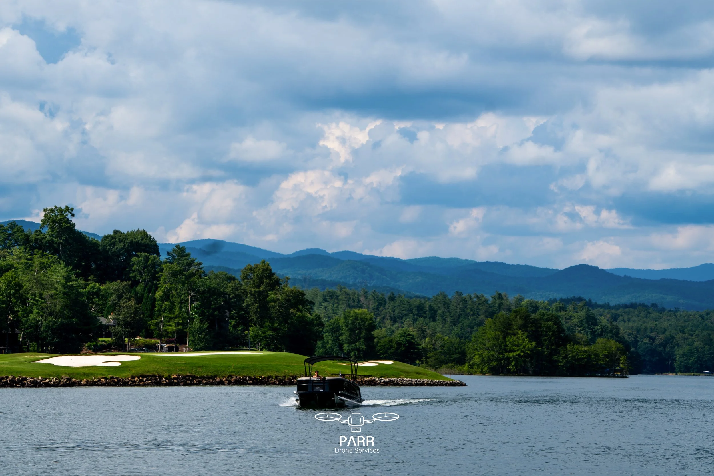 A boat is on a lake near a green golf course with sand traps, surrounded by trees and mountains under a cloudy sky.