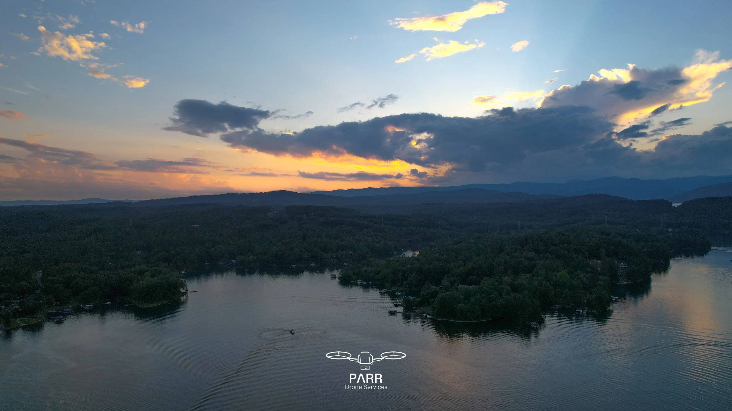 Aerial view of a lake surrounded by lush green trees with mountains in the background during sunset, with clouds in the sky.