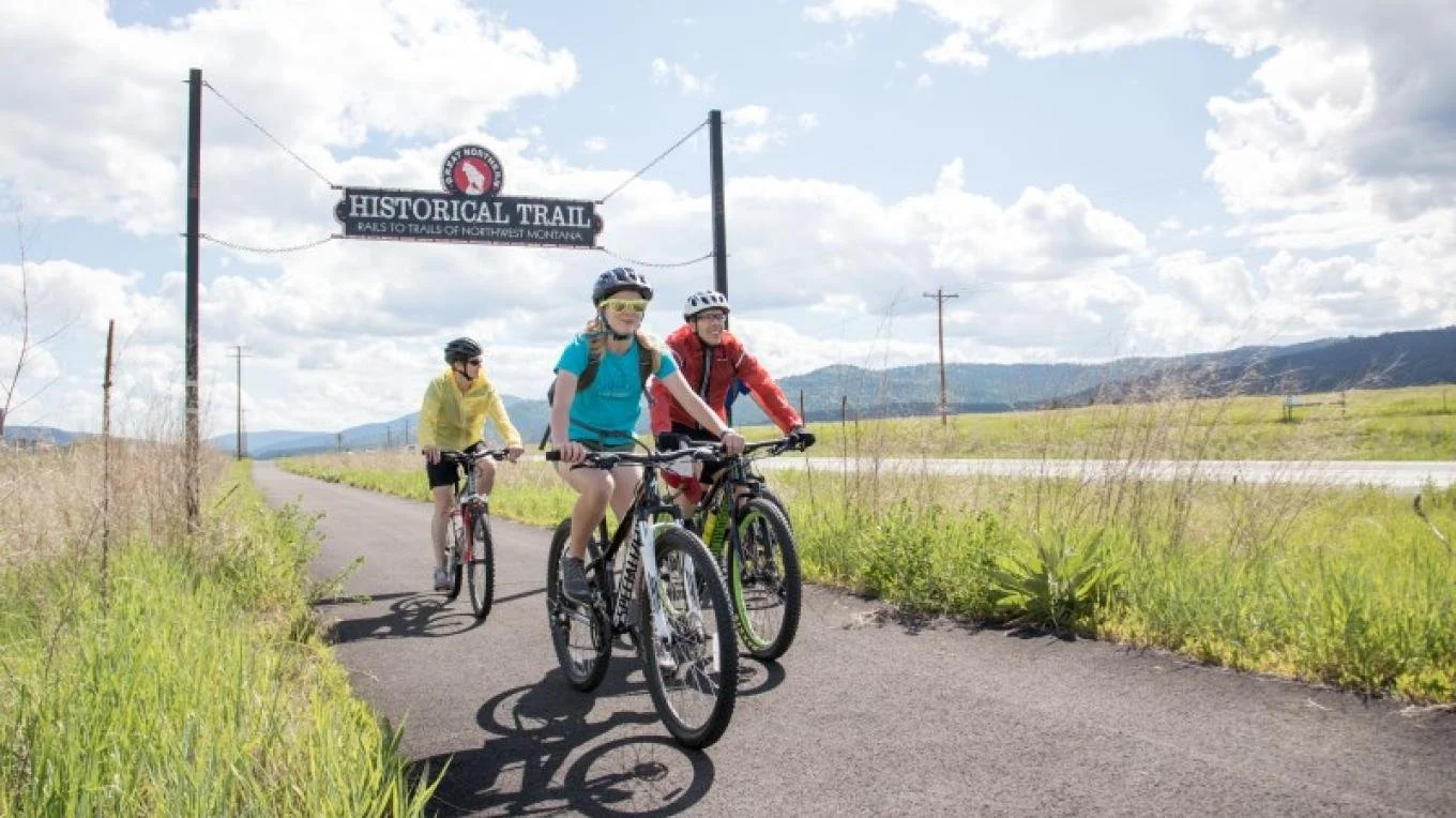 Three bicyclists ride on a sunny day on a paved rails to trails path.