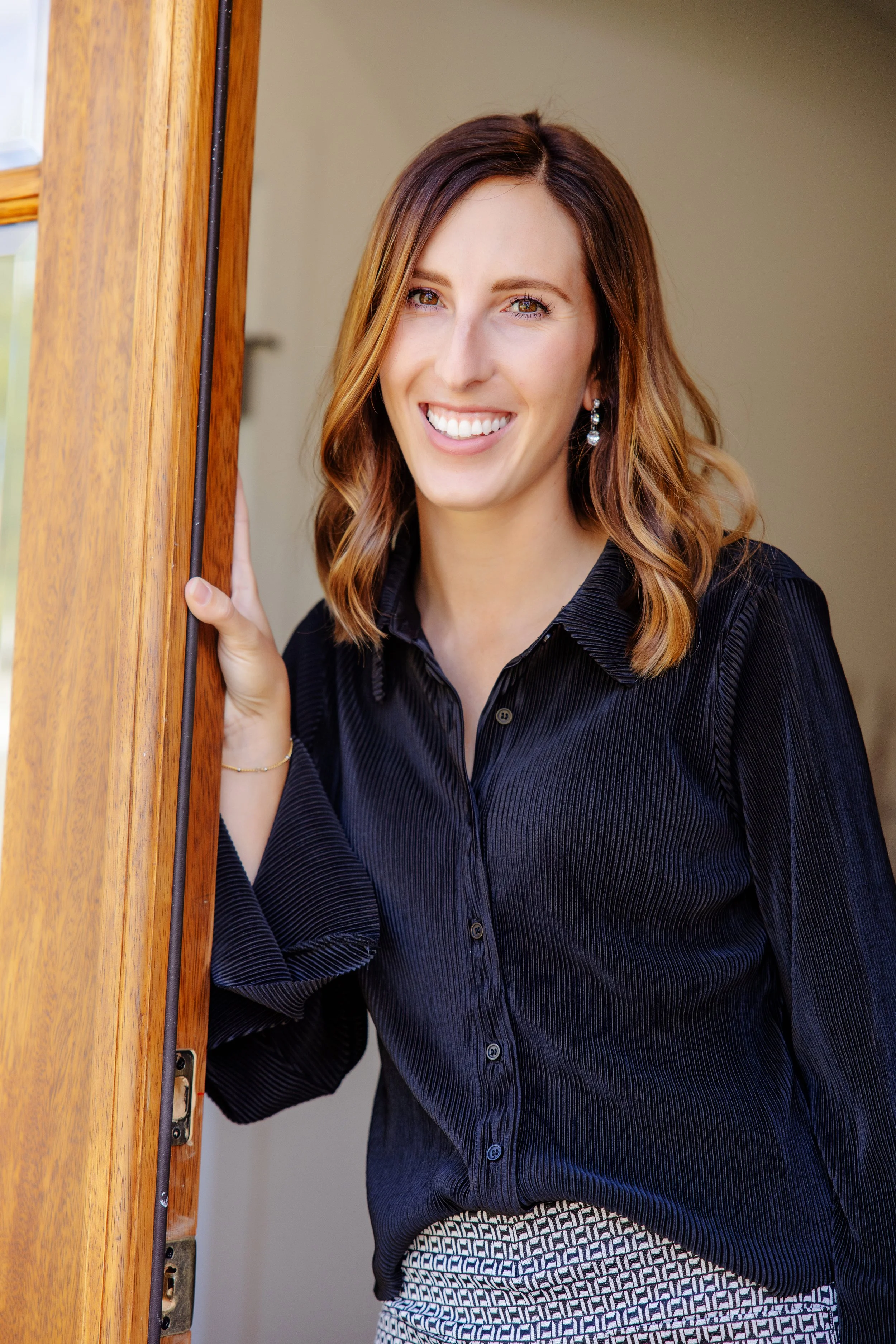 A smiling woman with wavy, shoulder-length hair standing by a doorway, wearing a black blouse and patterned skirt.