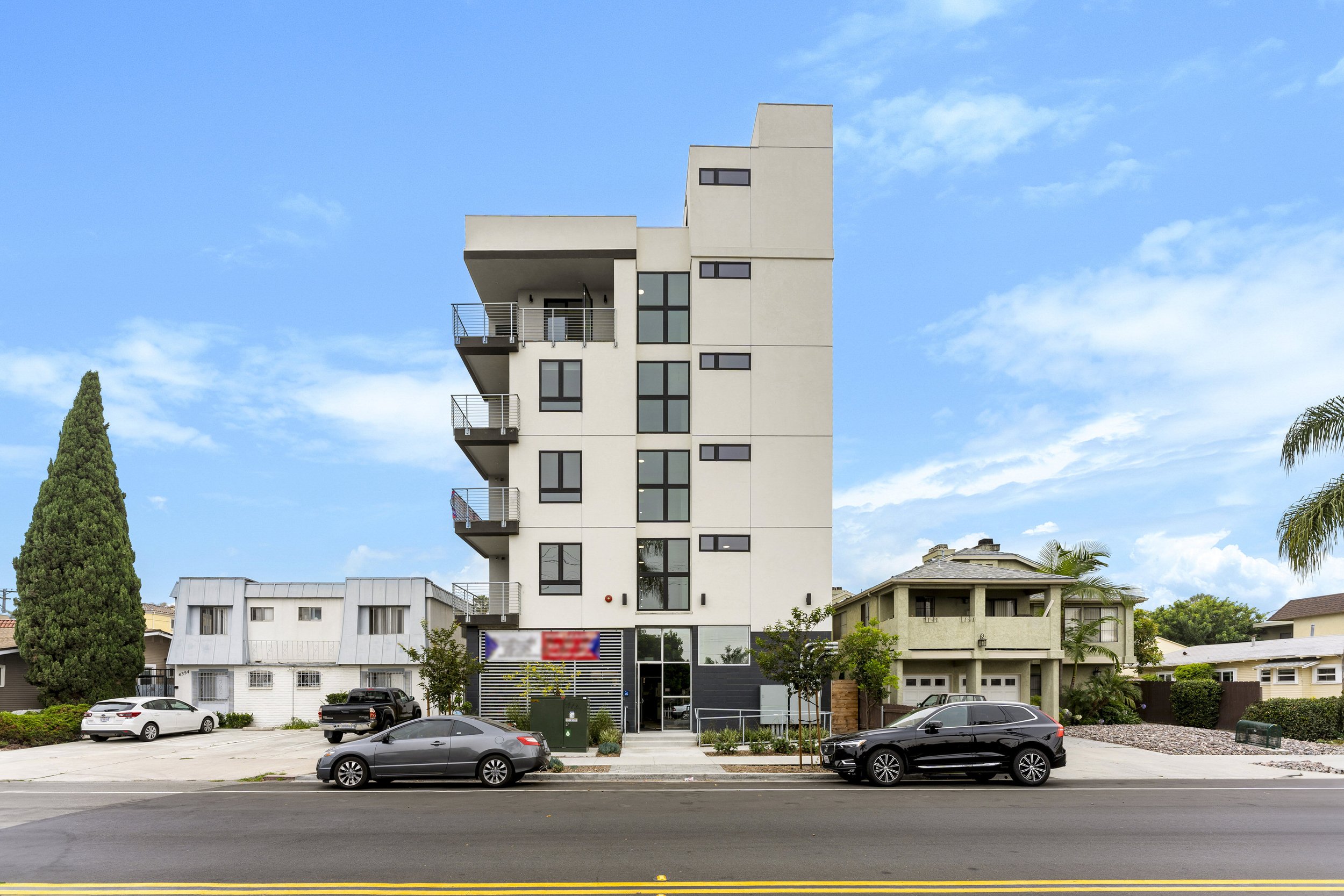 Open-concept living room and kitchen with white cabinets, stainless steel refrigerator, black kitchen island with wooden barstools, beige sofa with throw pillows and a blanket, abstract wall art, potted plant, and wooden flooring.