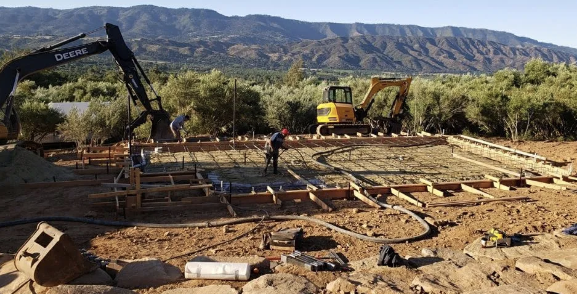Construction site with workers pouring concrete and scaffolding, surrounded by trees and mountains in the background.