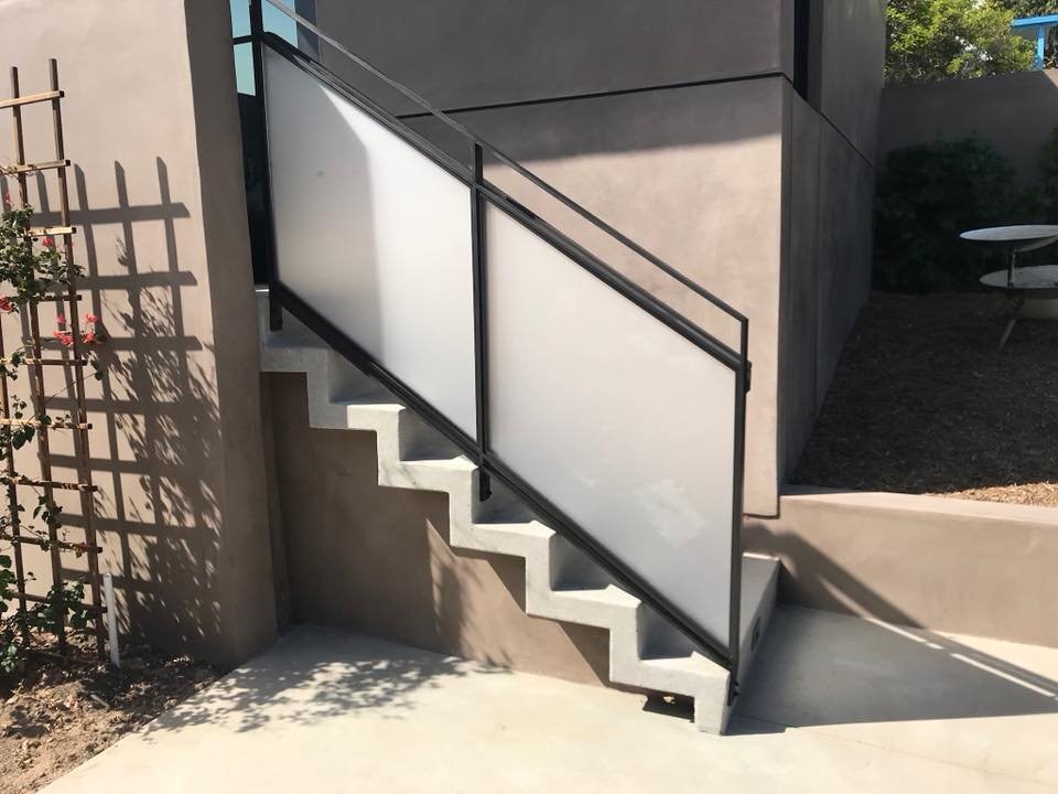 Concrete staircase with a black metal and frosted glass railing leading up to an outdoor area. There is a wooden trellis with climbing plants on the left and a round picnic table with benches on the right. Shadows are cast on the ground and wall.