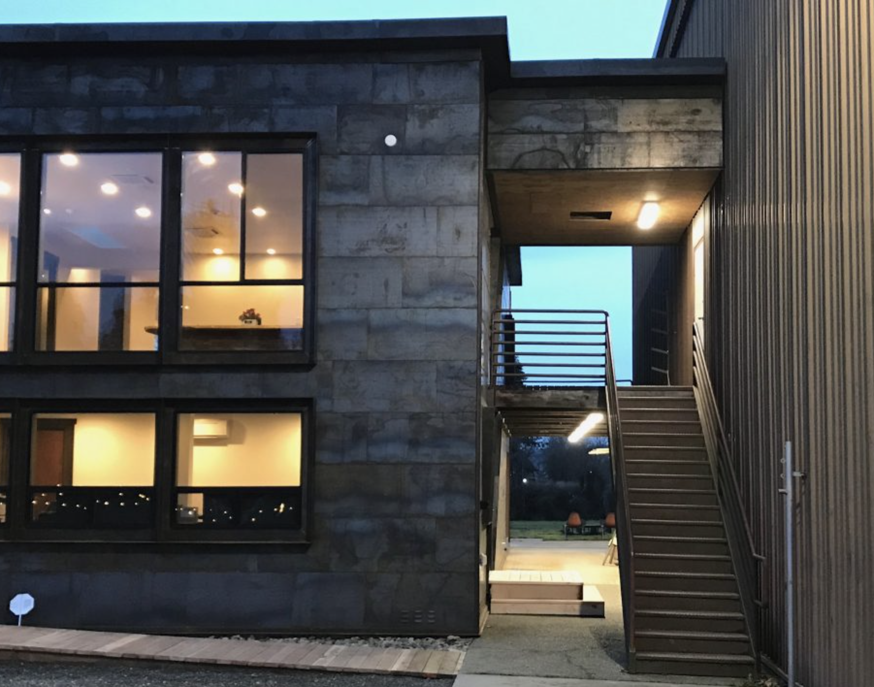 Exterior of a modern two-story building at dusk with large glass windows and a staircase leading to the upper level, illuminated by exterior lighting.