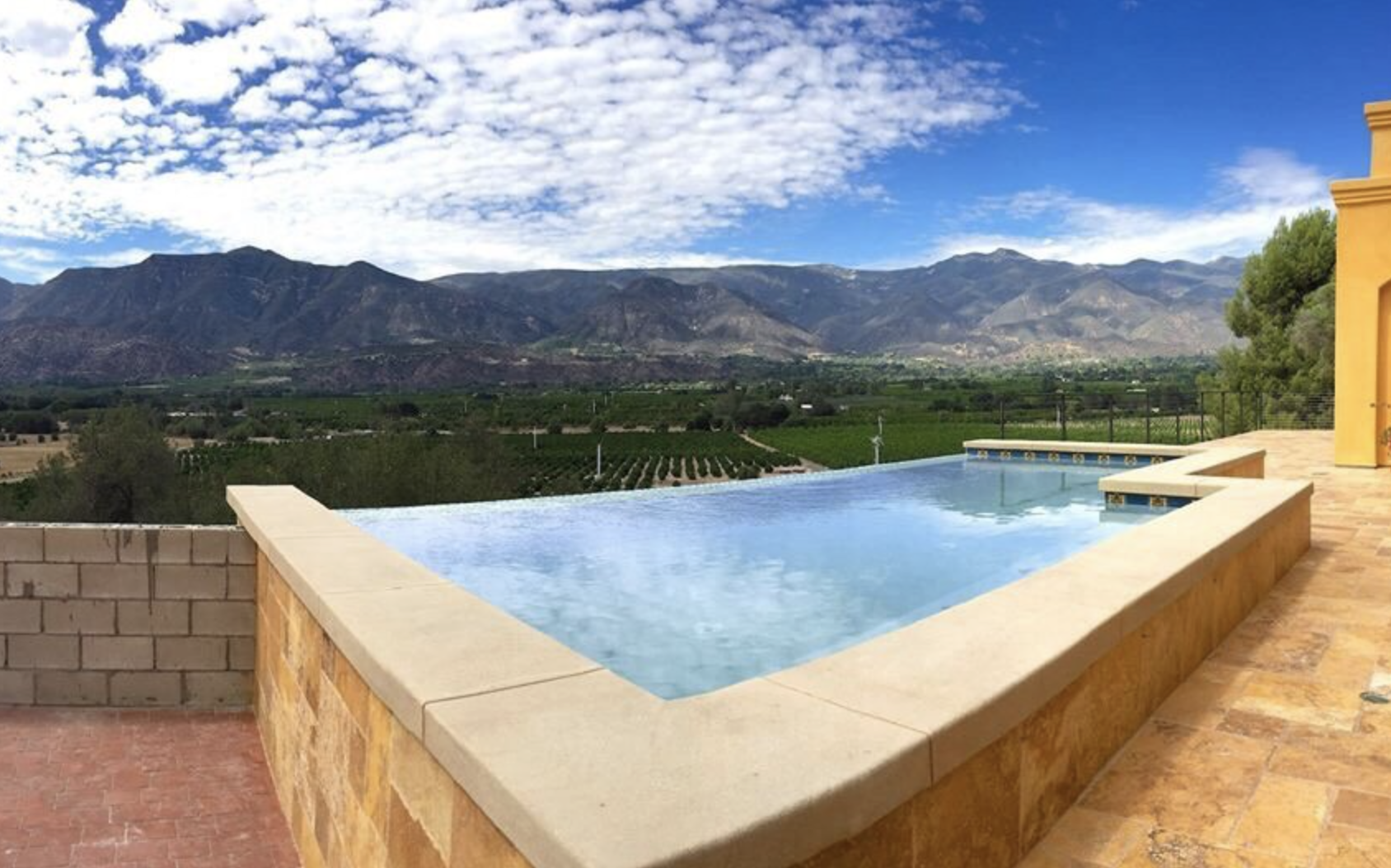 An infinity pool overlooking a mountain range with vineyards and a partly cloudy sky.