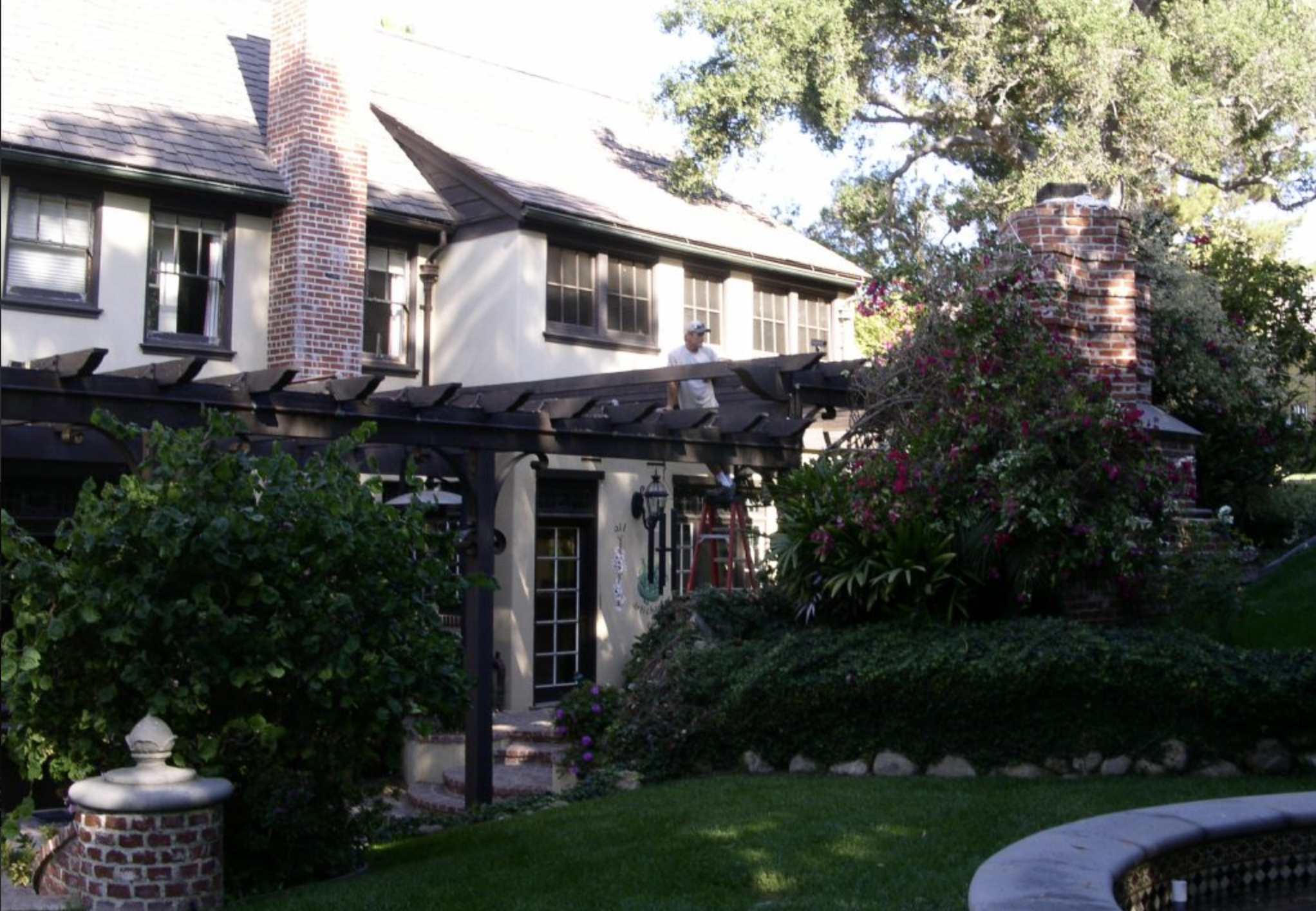 Man standing on roof of a house, working on the dark-tiled roof, with green bushes and flowering plants nearby in a well-maintained garden.