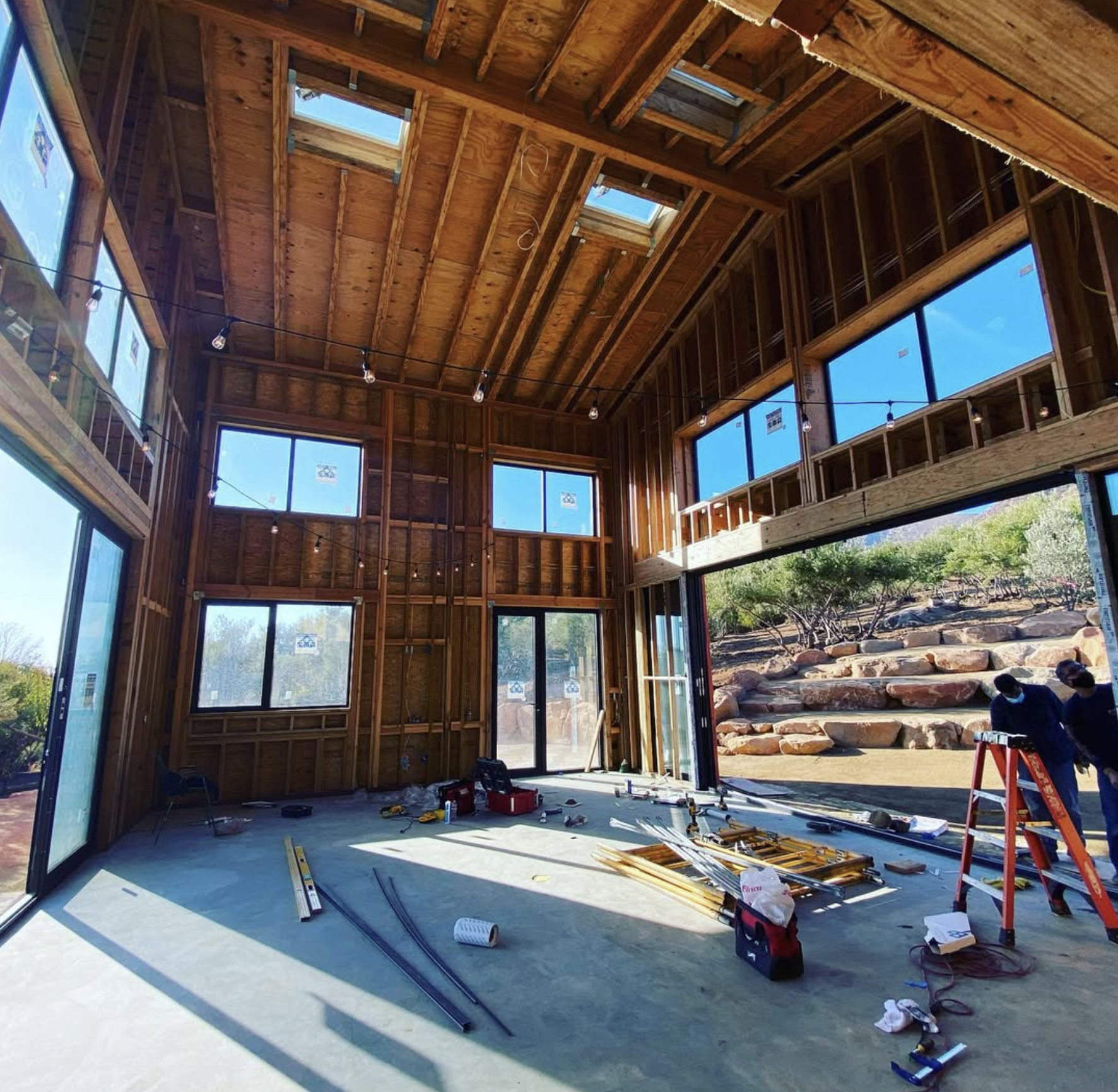 Construction site inside a wooden-framed building with large windows and a high ceiling, showing tools and equipment scattered on the floor, with two workers working near the entrance and outdoor landscape visible through the opening.