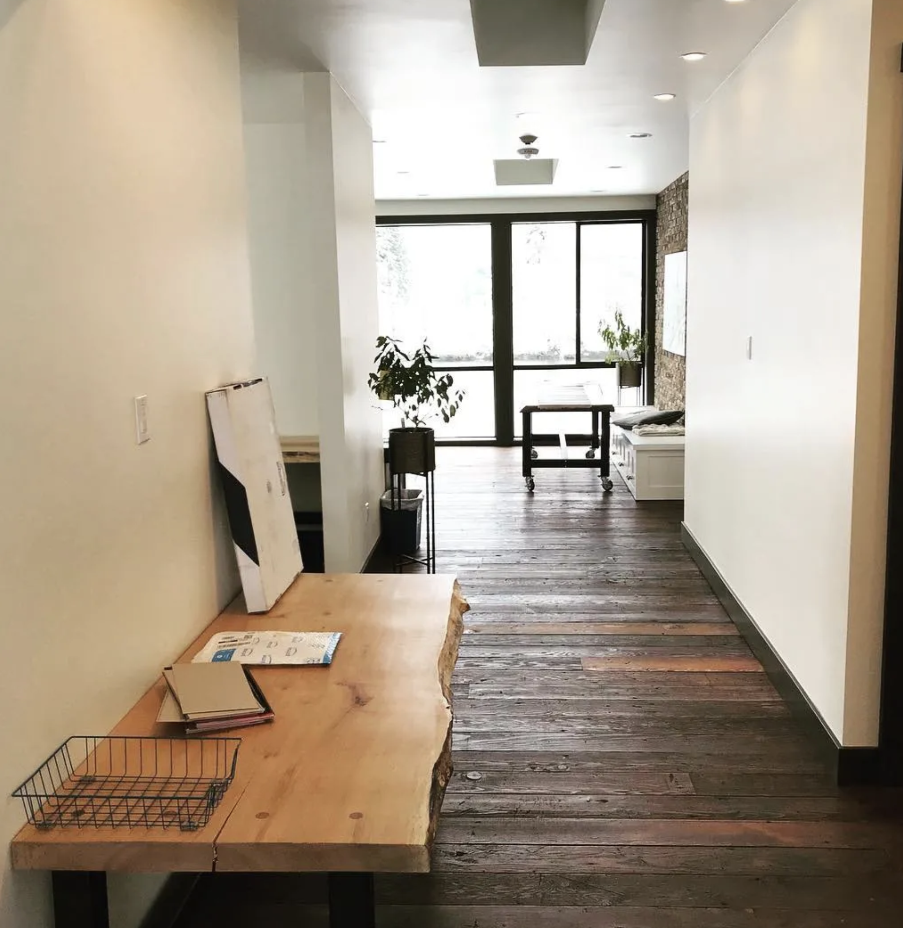 View of an interior hallway with wooden floors, a wooden table on the left, and glass doors at the end leading outside, with some potted plants near the doors.