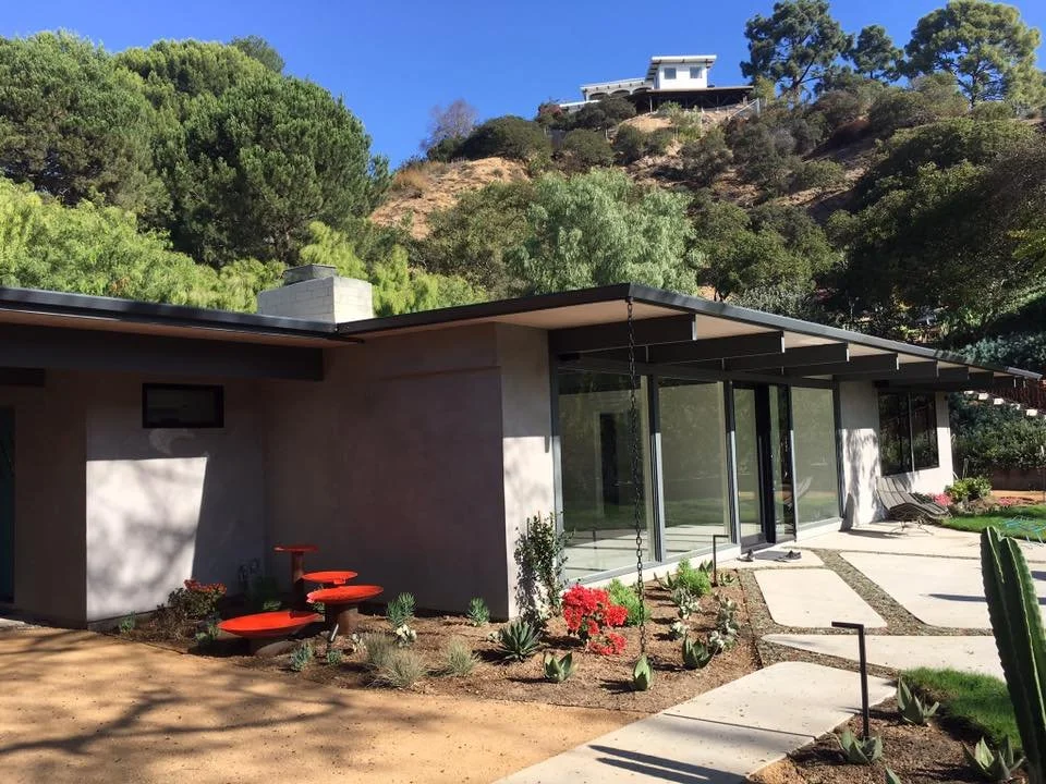 A modern house with a flat roof and large glass sliding doors, situated on a hillside with lush green trees and a clear blue sky. The yard has desert plants and a paved walkway.