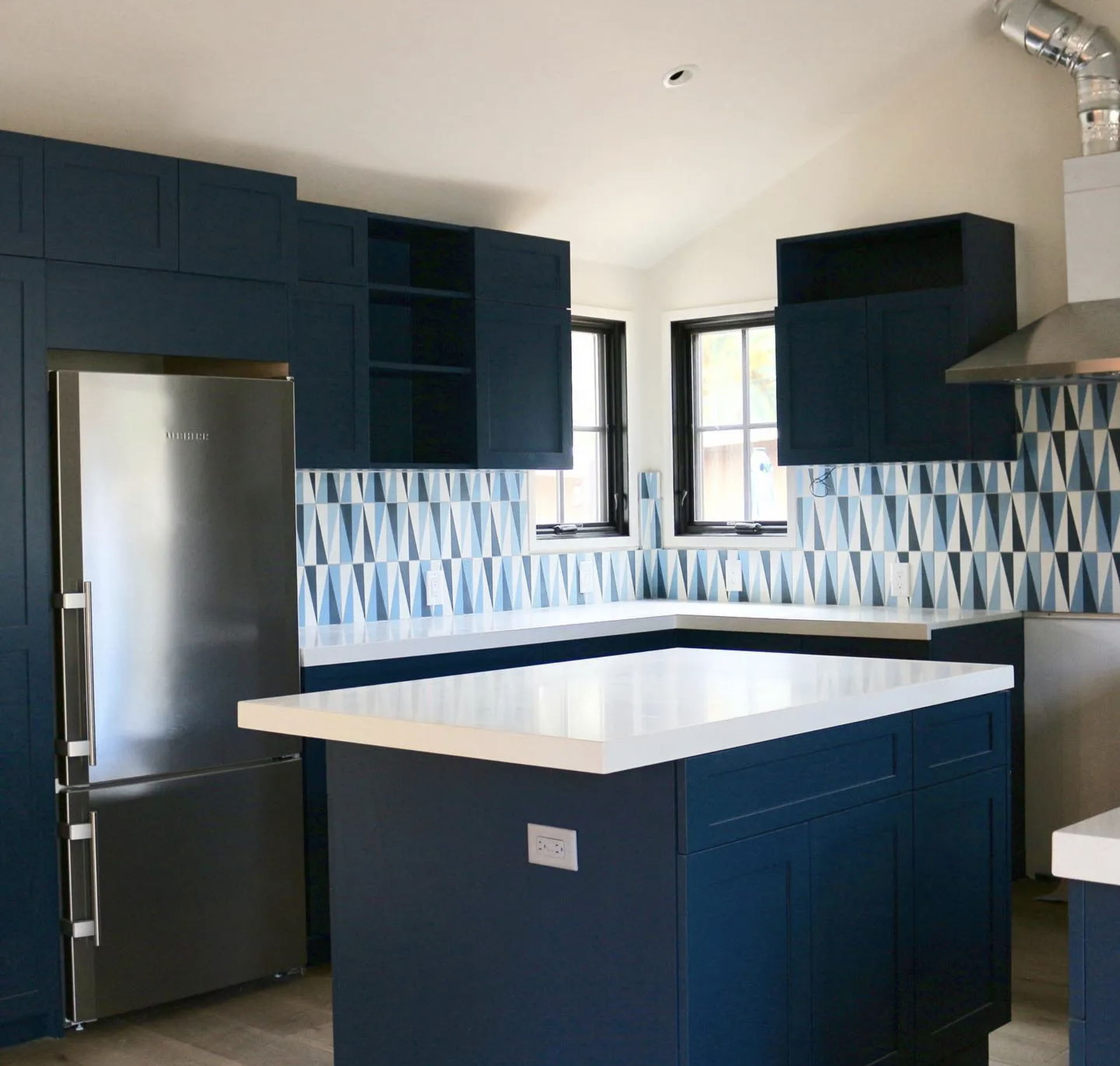 Modern kitchen with navy blue cabinets, geometric patterned blue and white tile backsplash, white countertop island, stainless steel refrigerator, and two black-framed windows.