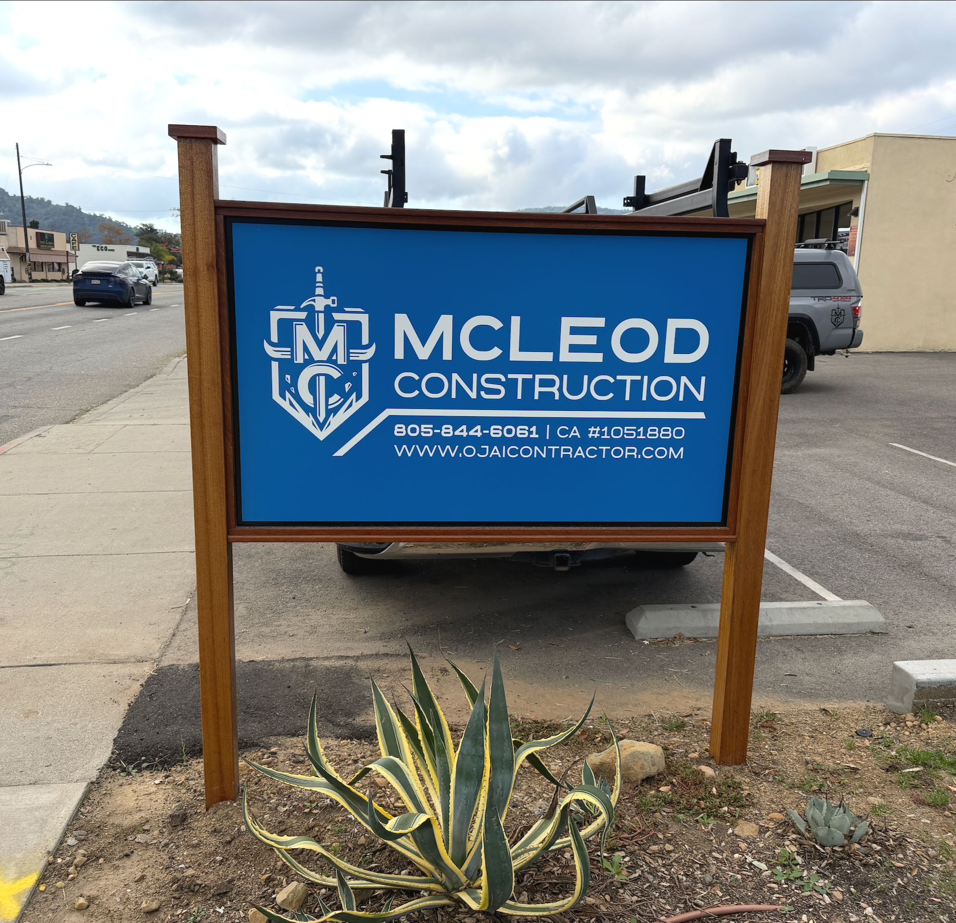 Blue sign for McLeod Construction with contact information, company logo, and website, placed on a sidewalk in a commercial area.