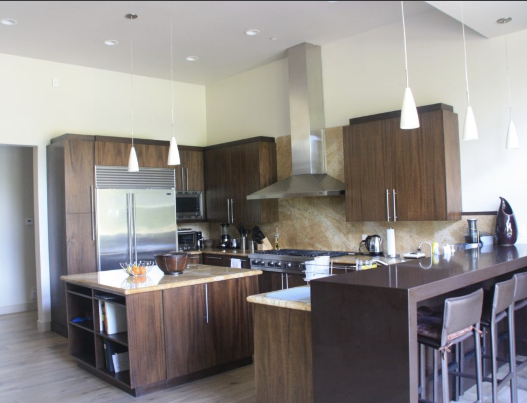 Modern kitchen with wooden cabinets, a stainless steel refrigerator, a kitchen island with a marble countertop, and a breakfast bar with three chairs, pendant lights, and various kitchen appliances