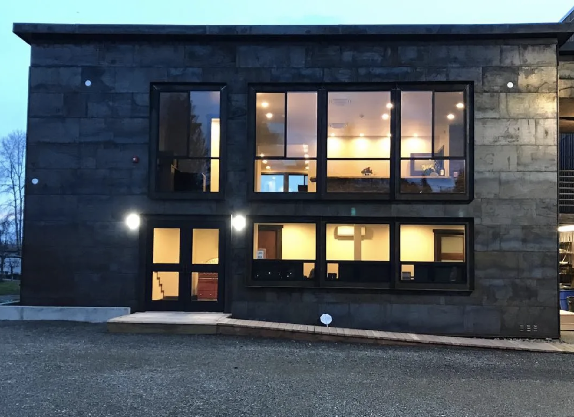 Modern two-story building with large windows, black exterior, illuminated interior, and gravel driveway.
