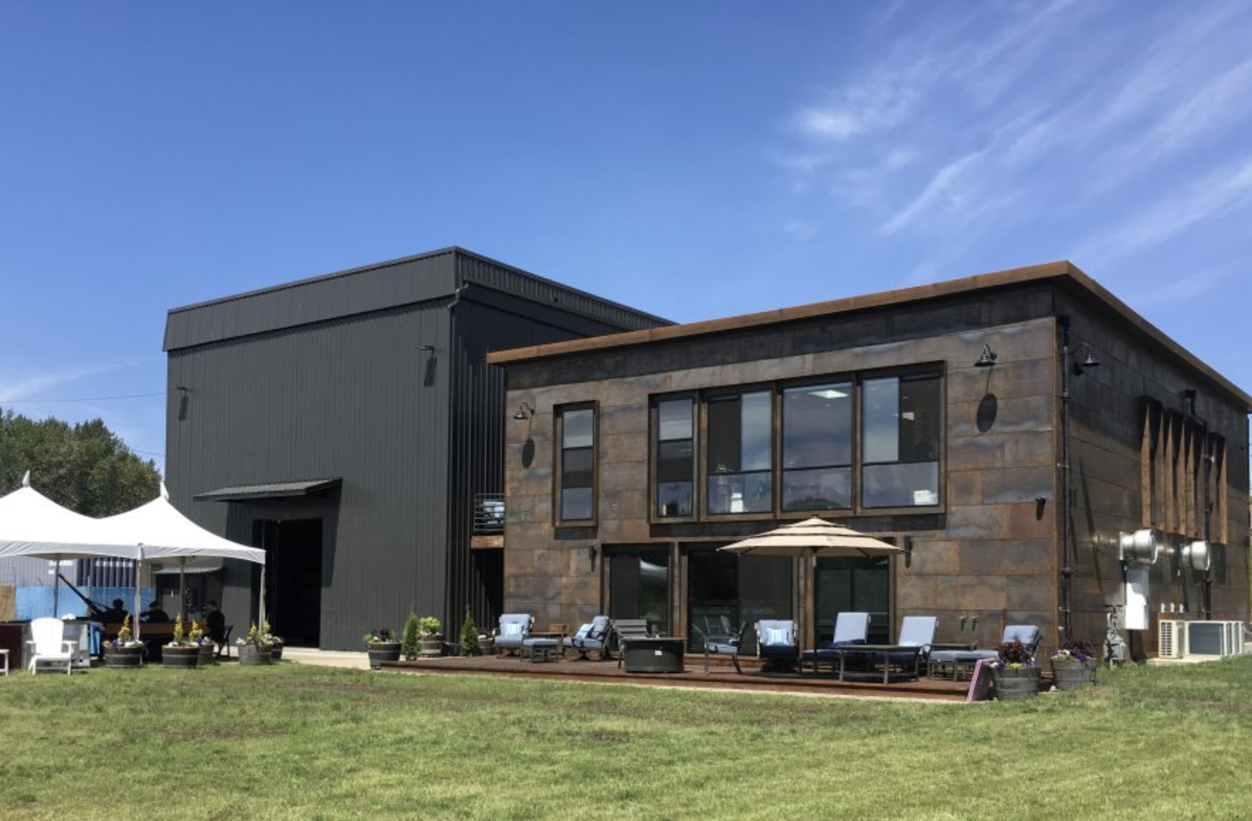 Exterior of a modern building with a patio, seating, and umbrellas, set on a grassy area under a blue sky.