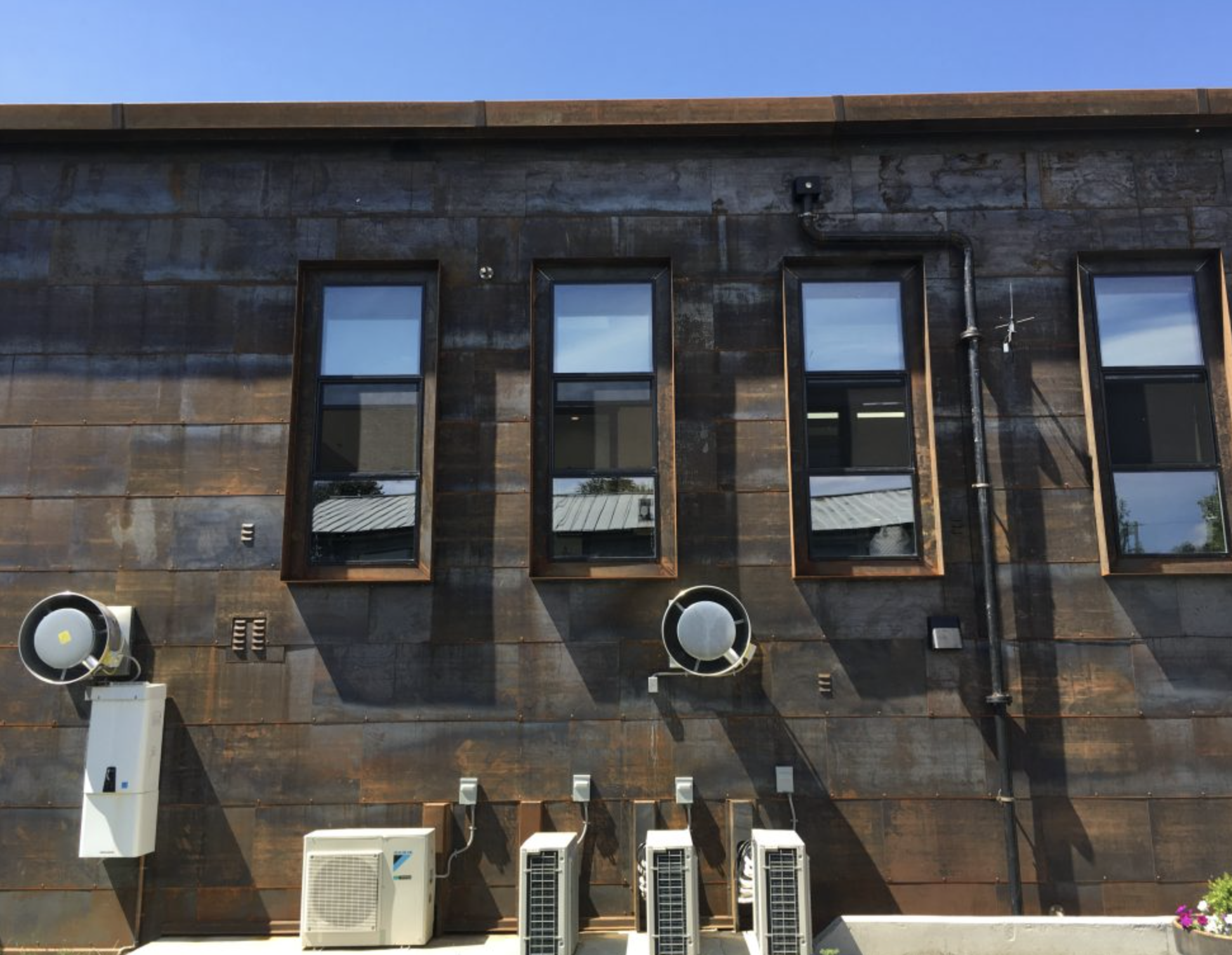 The exterior of a modern building with four tall, narrow windows and various air conditioning units and vents on the wall, under a clear blue sky.