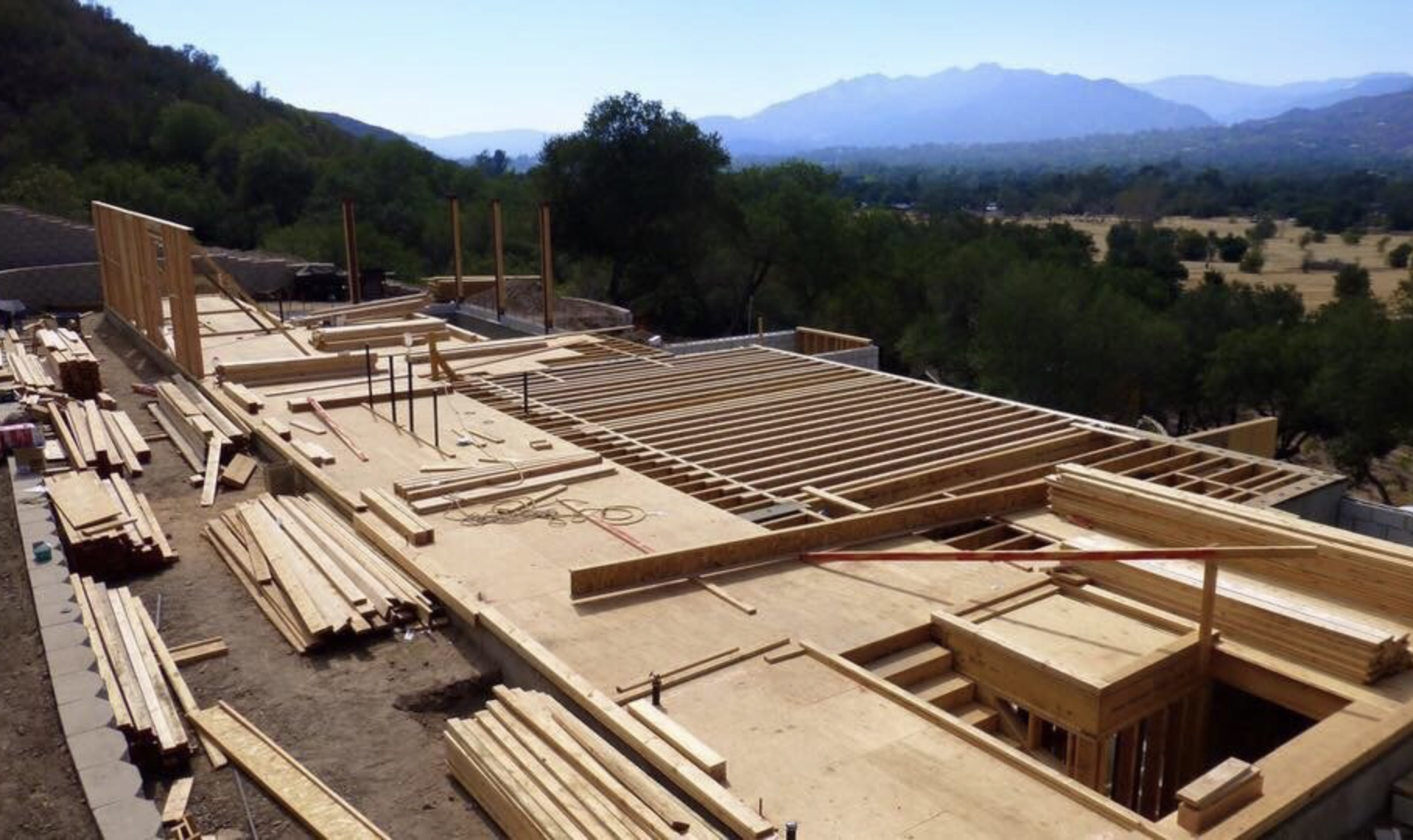Construction site with wooden framework for a building, surrounded by trees and mountains in the background.