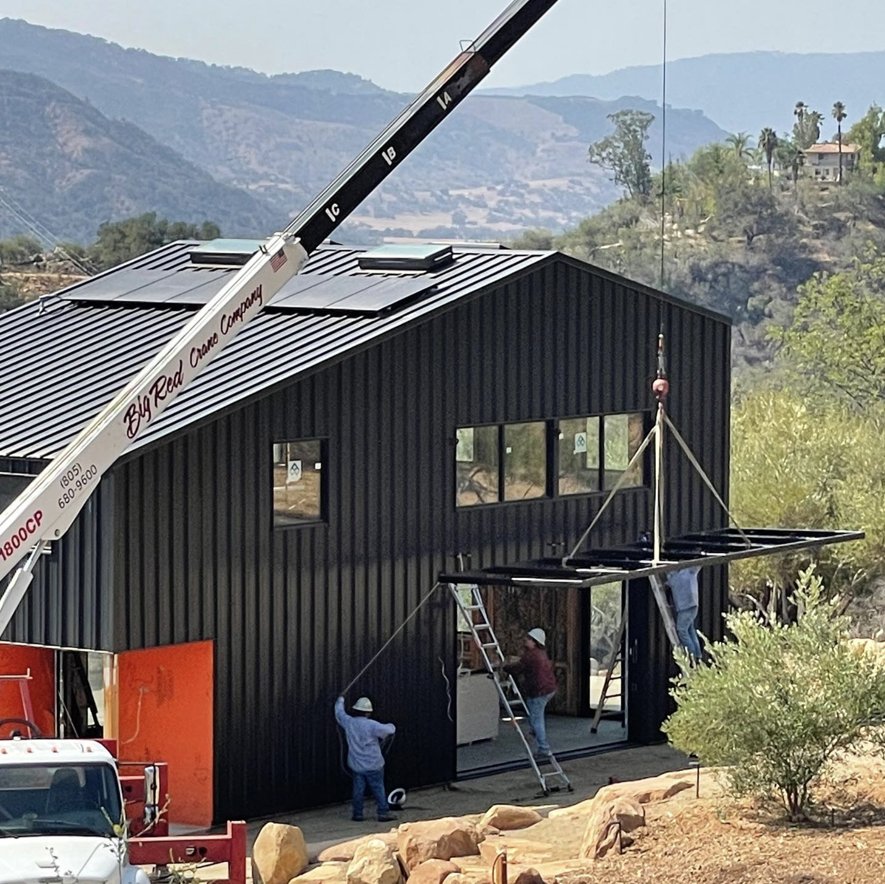 Construction workers installing a black metal building structure with a crane on a hillside with mountains and trees in the background.