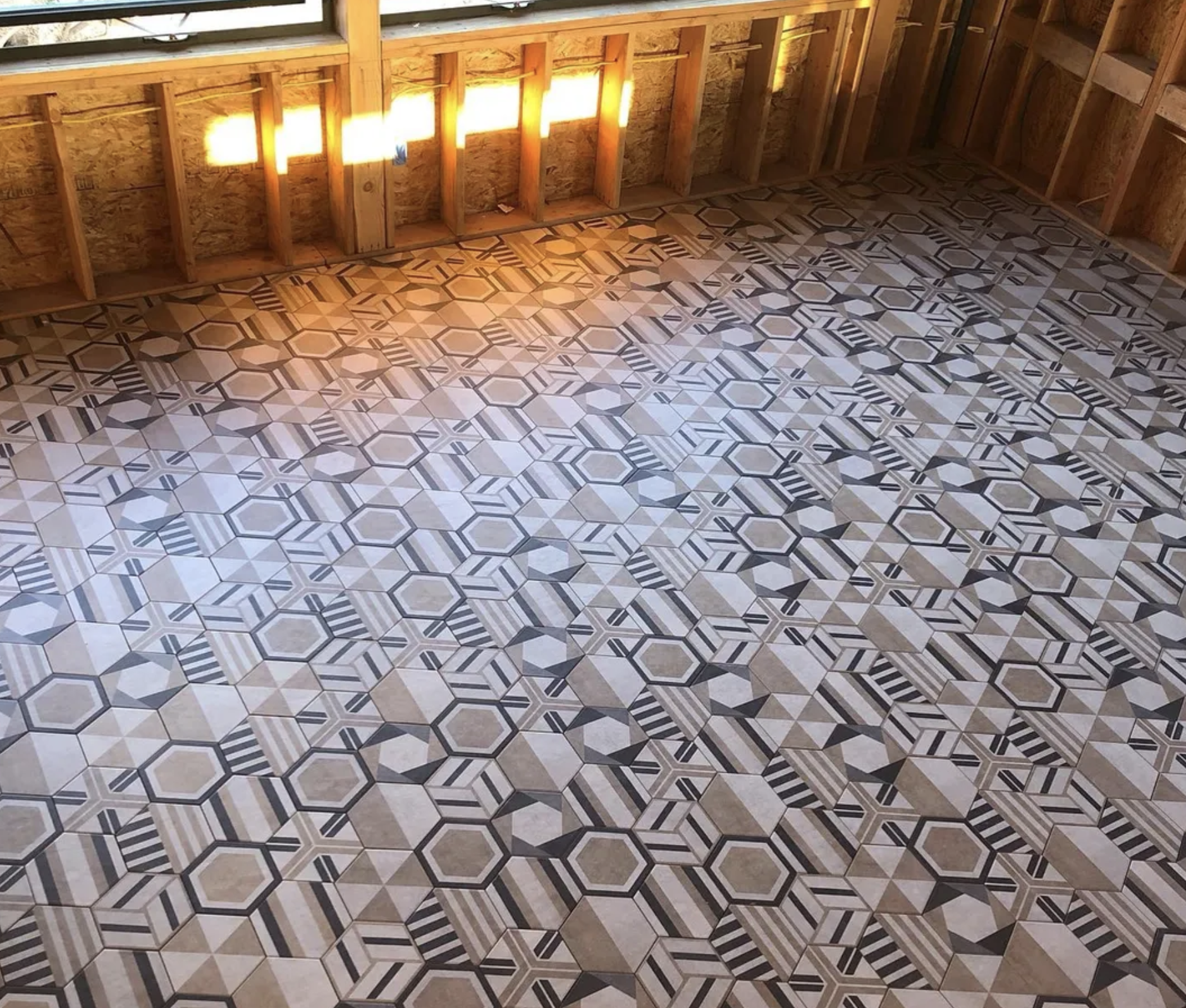 An unfinished interior space with a decorative tile floor featuring a geometric pattern in shades of tan, cream, and black. The room has exposed wooden framing and plywood walls, with sunlight shining through a gap in the wall.