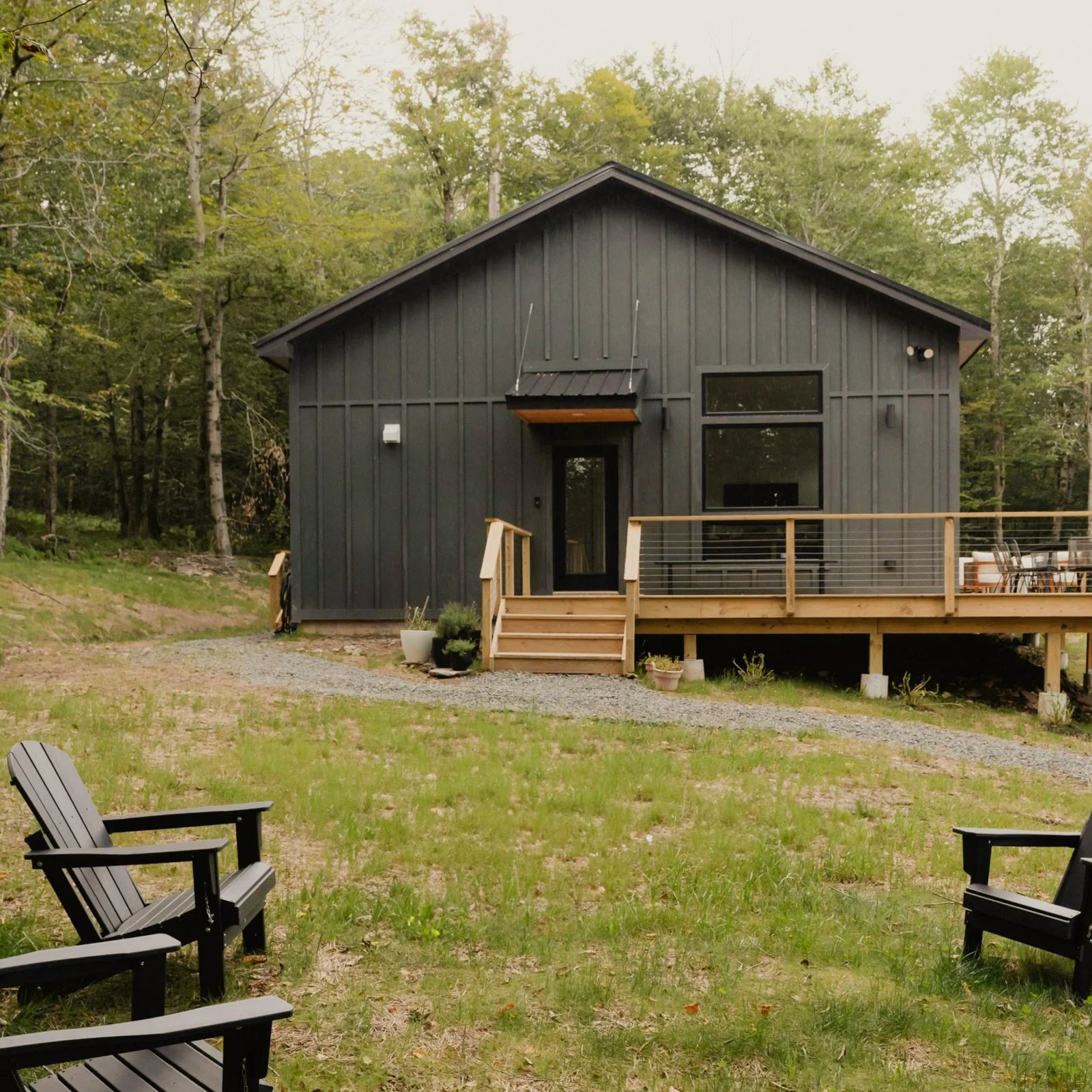 Modern black house with wooden deck and steps, surrounded by green grass and trees.