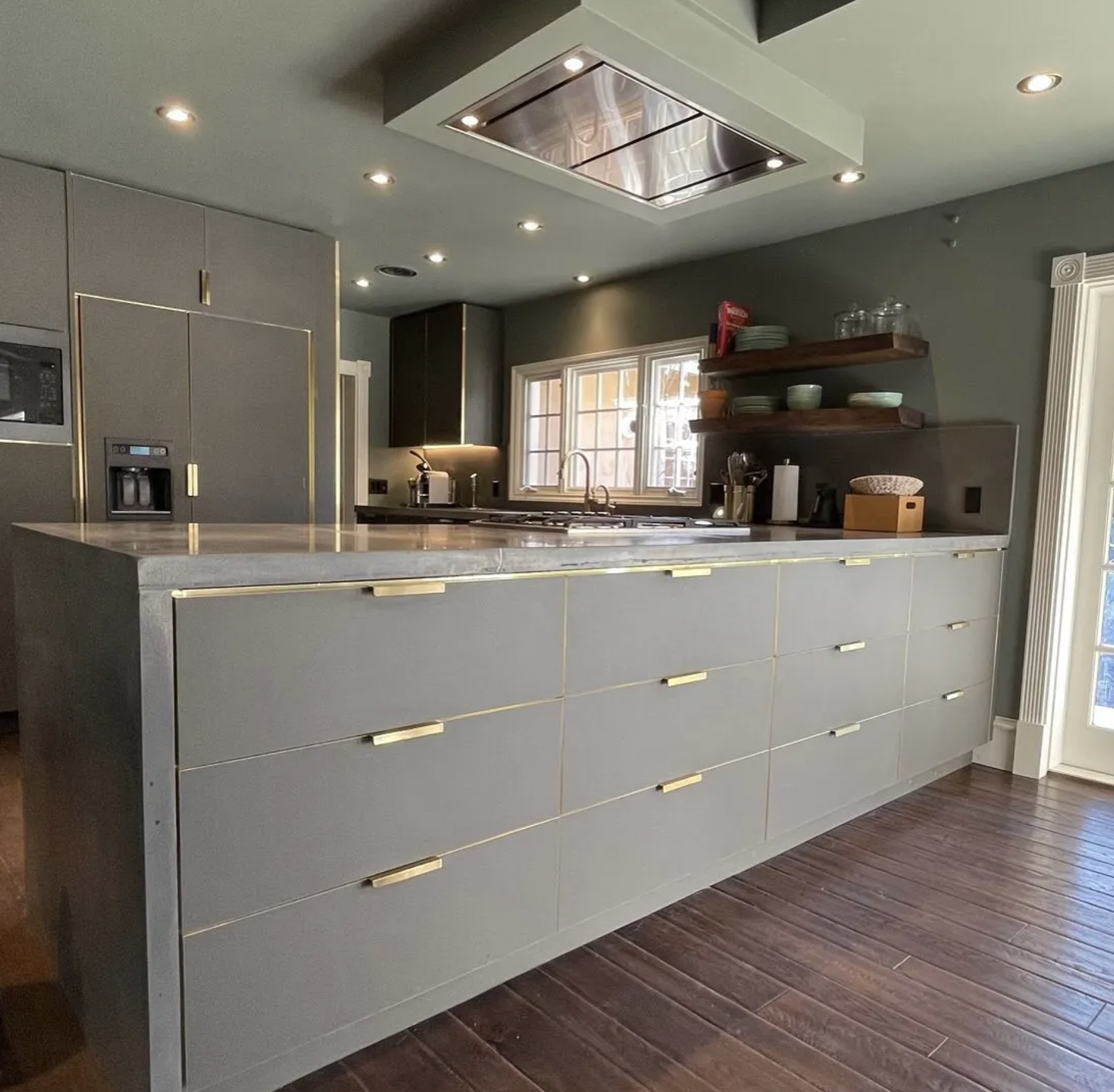 Modern kitchen with grey cabinets, gold handles, marble countertop, window above sink, open wooden shelves with dishes, and dark wood flooring.