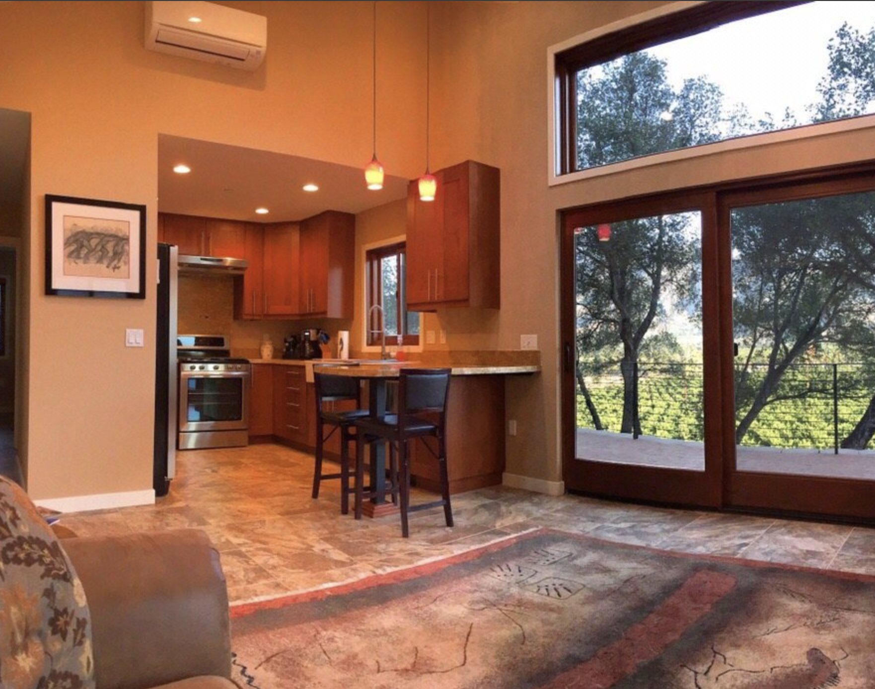 Interior view of a kitchen and living area with large windows overlooking trees, wooden cabinetry, a small breakfast bar with two chairs, and a patterned rug on a tiled floor.
