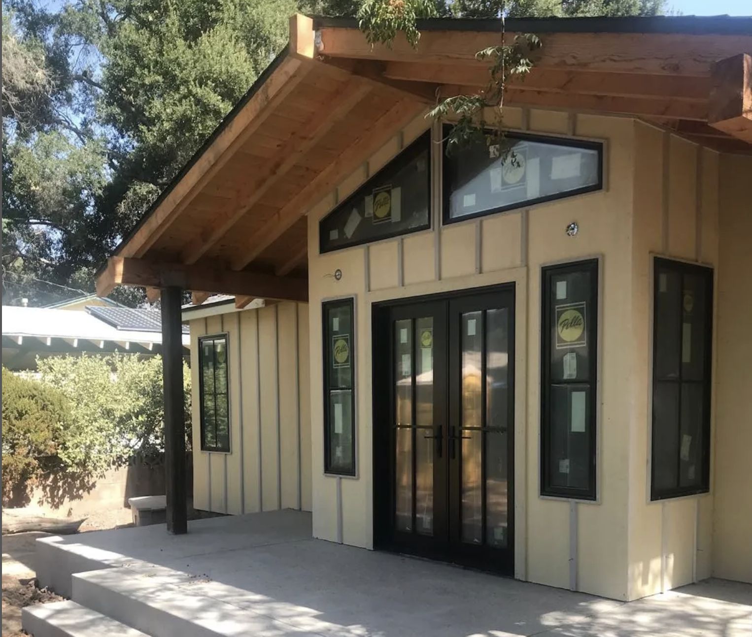 New house with black framed windows and double glass doors, wood siding, concrete steps, and a sloped wooden roof supported by a black post, surrounded by trees.