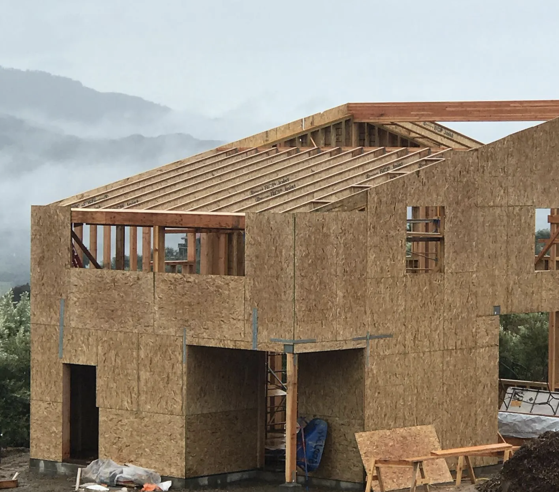 Construction of a two-story wooden house with plywood walls and roof framework on a cloudy day.