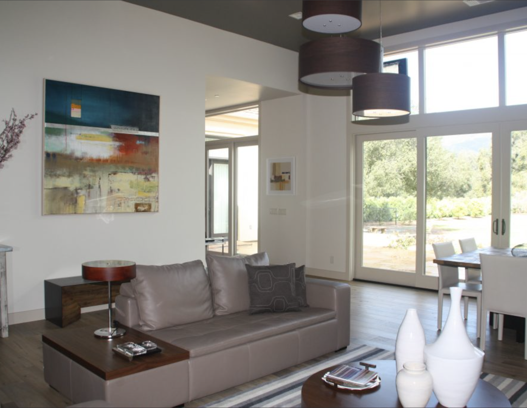Living room with beige sofa, abstract paintings, wooden coffee table with white vases, and large sliding glass doors showing outdoor greenery.