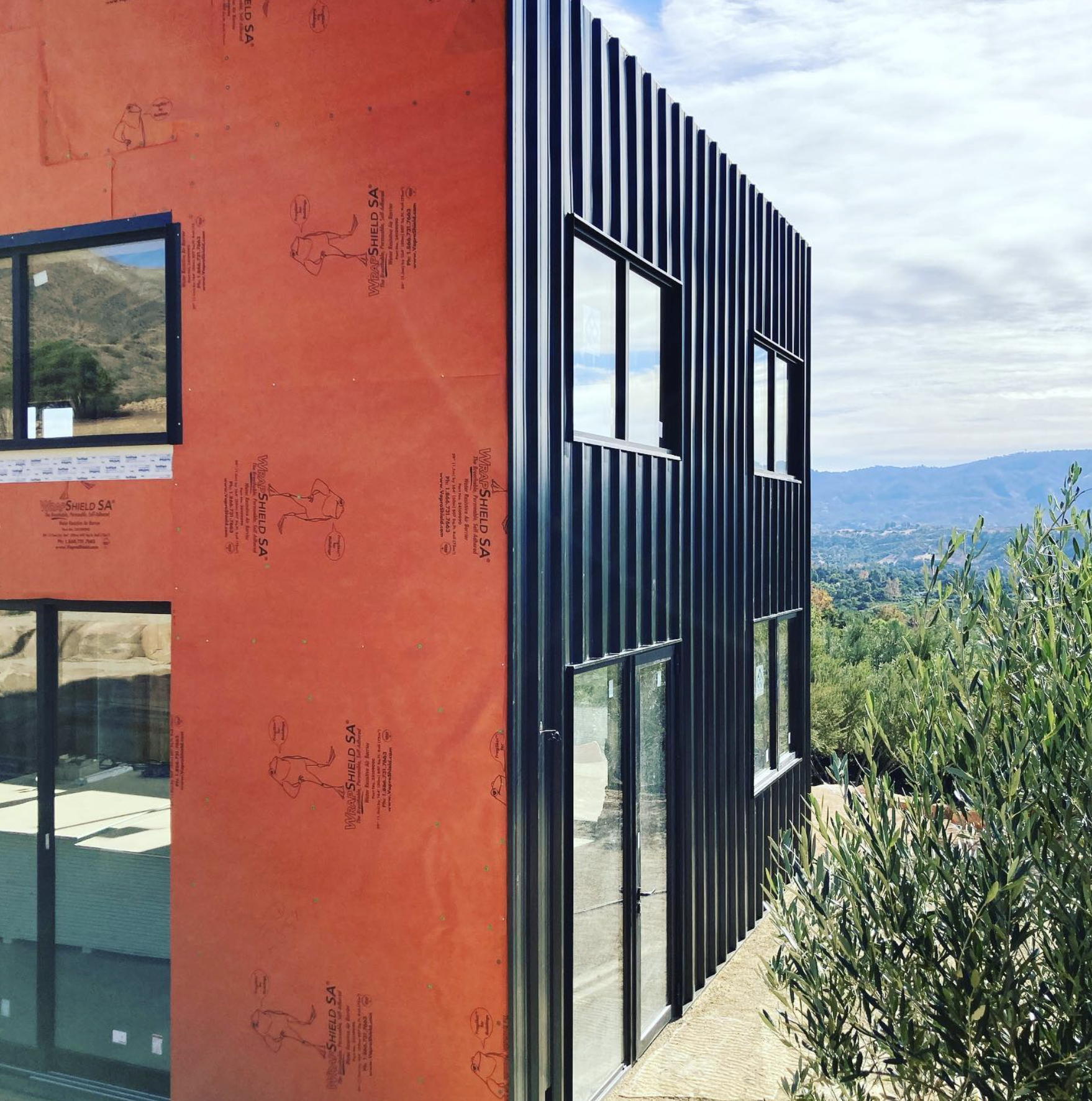 Partially constructed building with black metal siding and orange insulation panel, overlooking a scenic landscape with trees and hills.