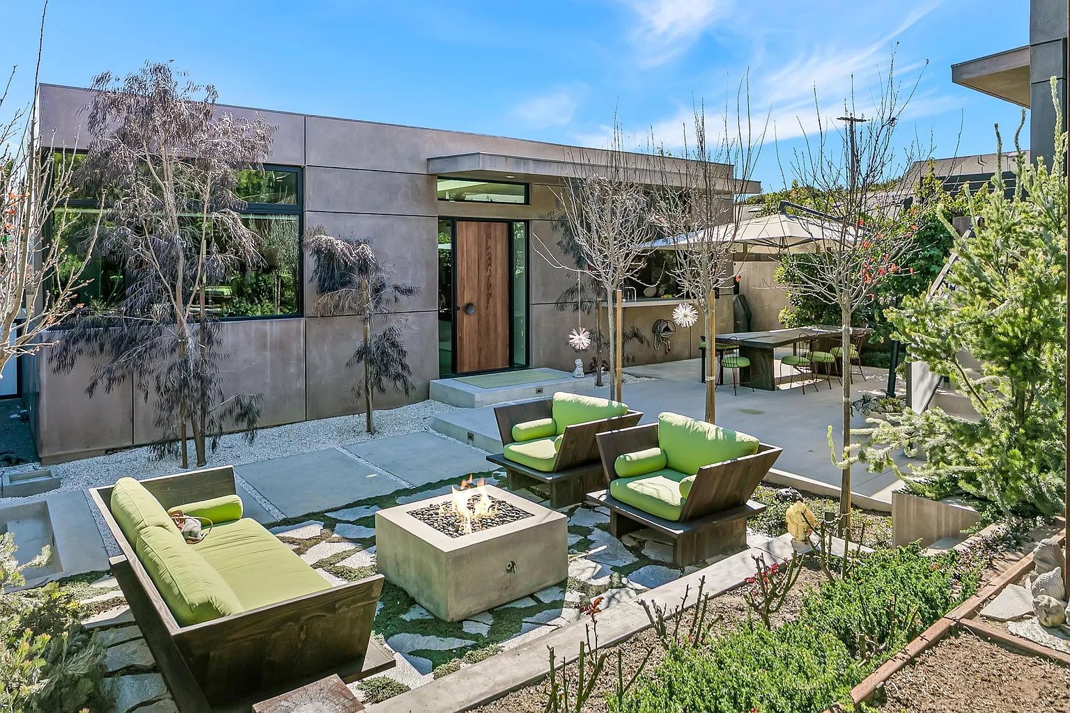 Modern backyard with outdoor seating, fire pit, trees, and a concrete patio next to a contemporary house with large windows and a wooden door.