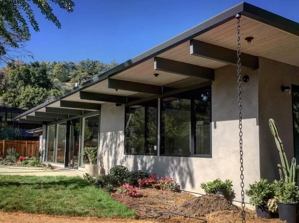 Modern house exterior with large windows, landscaped garden, and a wooden fence, under a clear blue sky.