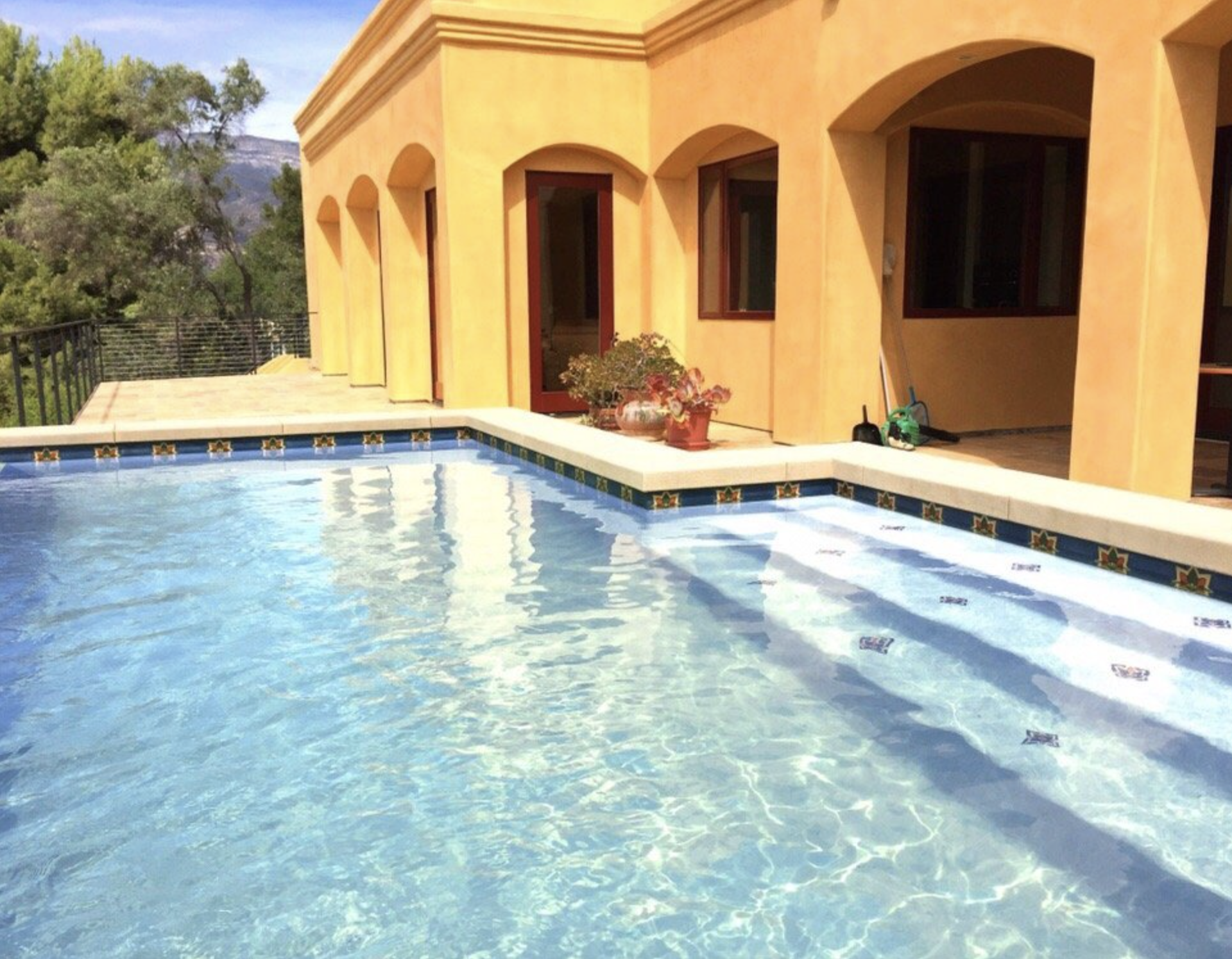 A swimming pool with clear water beside a yellow stucco house with arched windows and a door, surrounded by greenery and mountains in the background.
