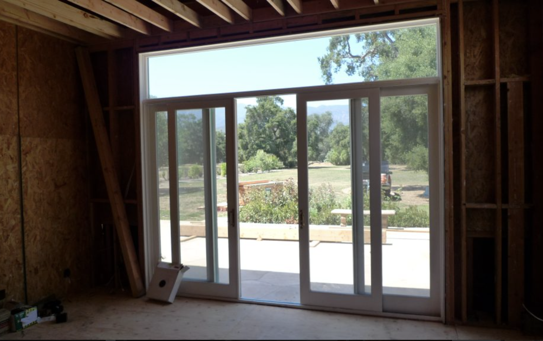 Interior view of a house under construction with large sliding glass doors opening to a backyard with trees and a grassy area.