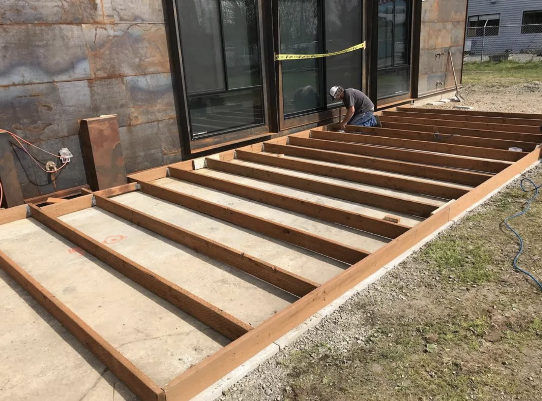 Construction worker kneeling and working on a wooden deck framing outside a building with large glass windows, with construction tape and tools around.