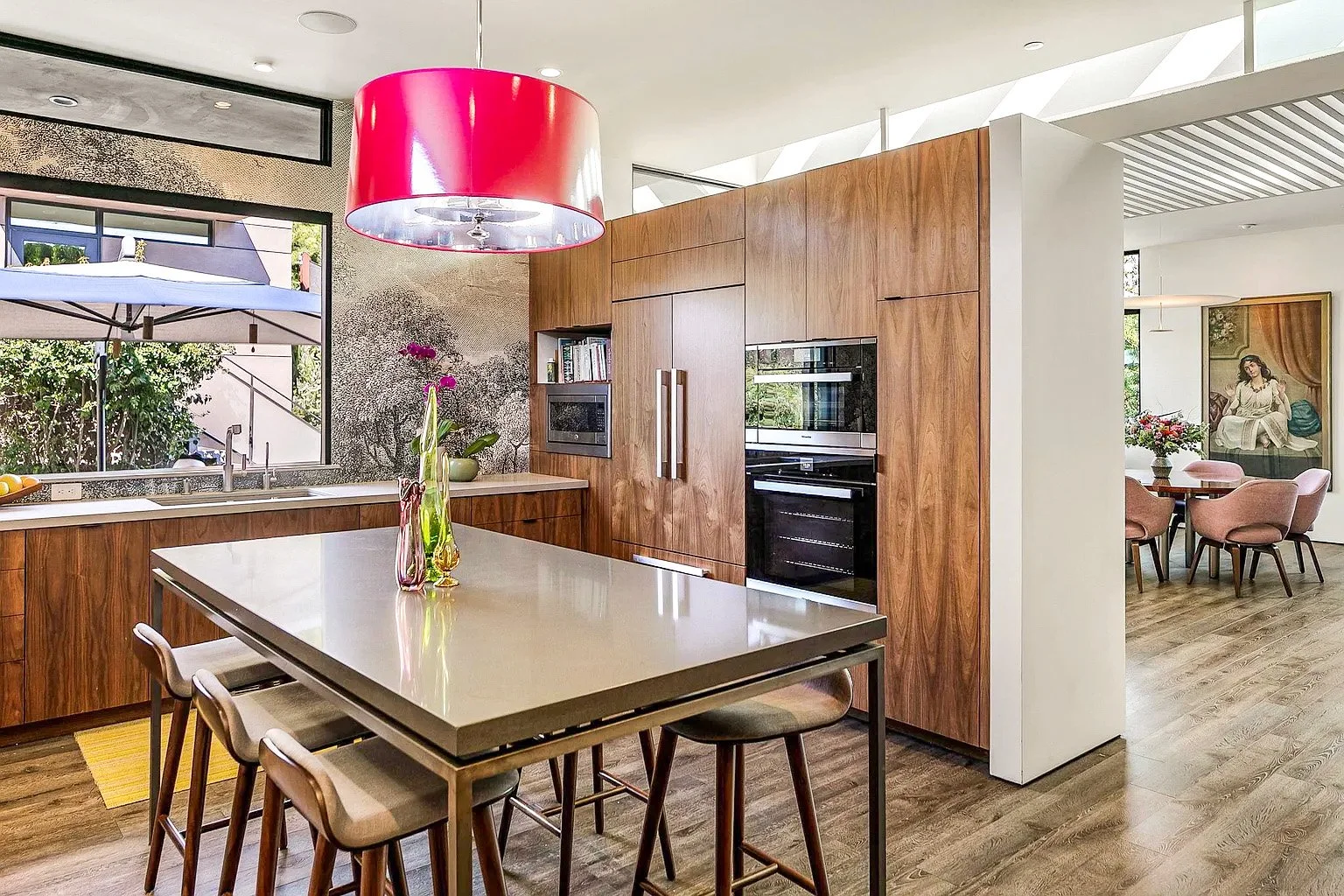 Modern kitchen with wooden cabinets, a white countertop, and a central island with a beige top. A large red pendant light hangs over the island. There are three barstools around the island. The kitchen has a window overlooking an outdoor patio with a parasol. To the right, there is a dining area with a round table, pink chairs, a vase of flowers, and a wall with artwork of a woman sitting.