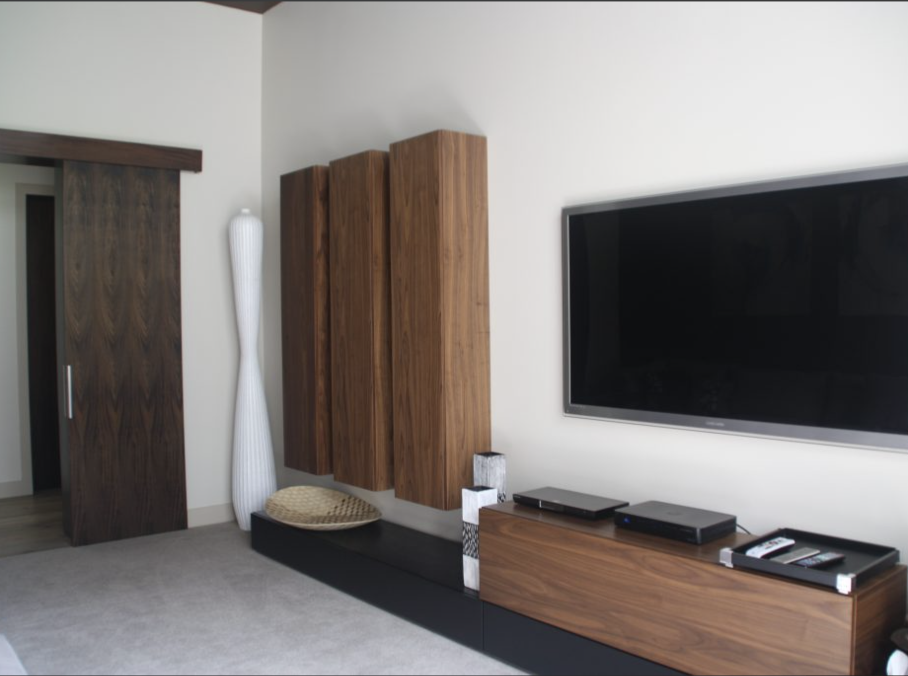 Modern living room with a flat-screen TV mounted on the wall, a wooden media console with electronic devices, wooden wall panels, a decorative white floor sculpture, a woven basket, and a dark wooden sliding door.