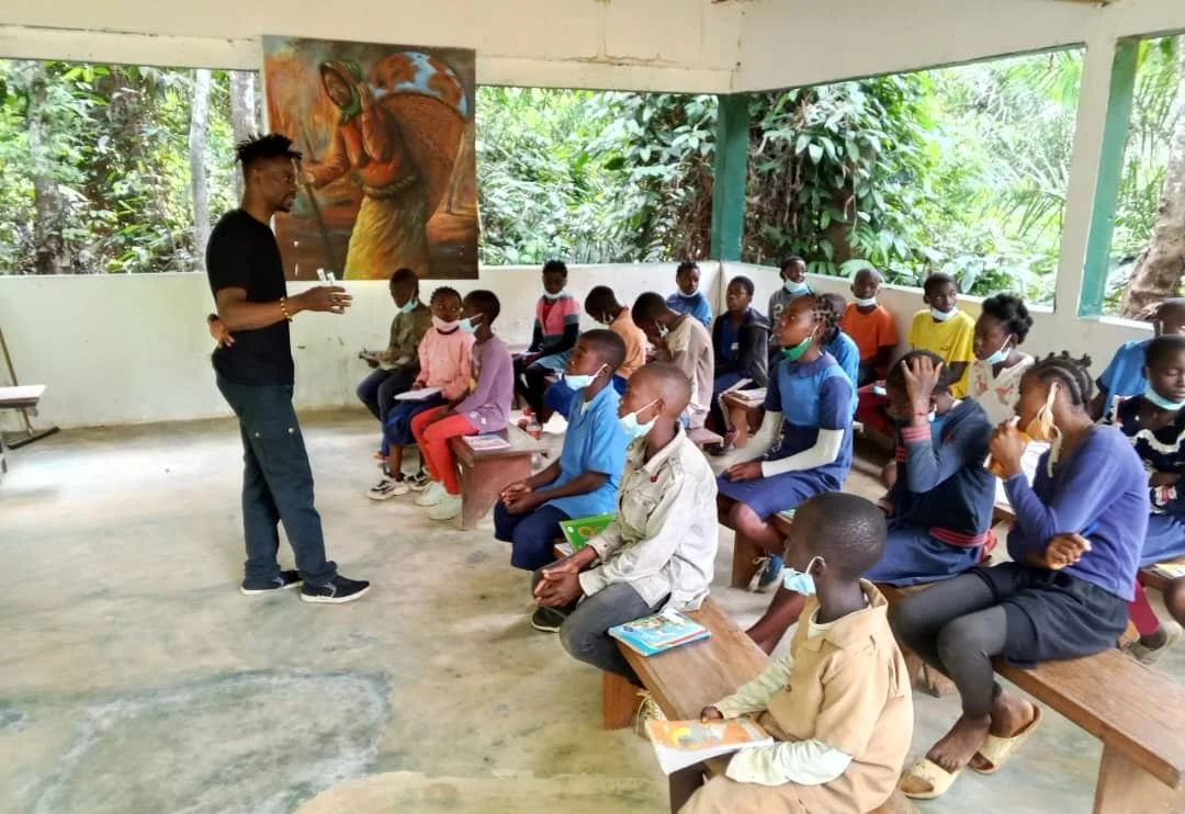 A classroom with children sitting on benches, wearing face masks, and a teacher standing in front of them. The classroom has open sides with greenery outside and wall art of an elephant.