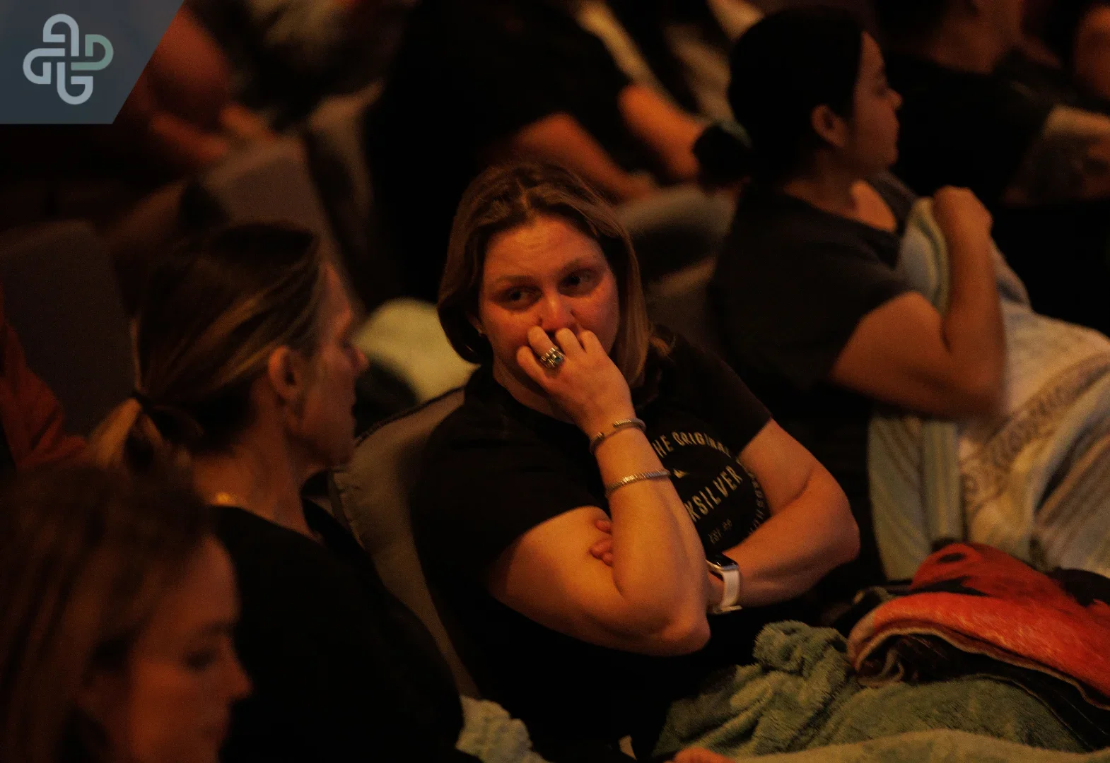 In a dimly lit audience, a woman covers her mouth and watches the stage with concern, surrounded by seated spectators; the mood is tense and absorbed.