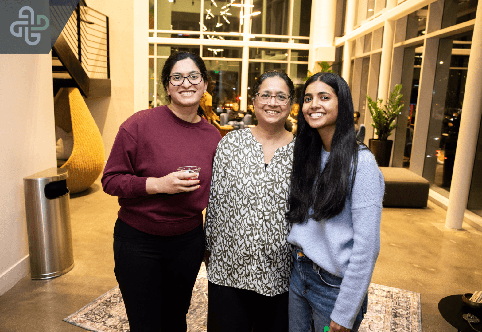Three smiling women stand together in a brightly lit modern office lobby or event space during a community gathering hosted by The Den Meditation.