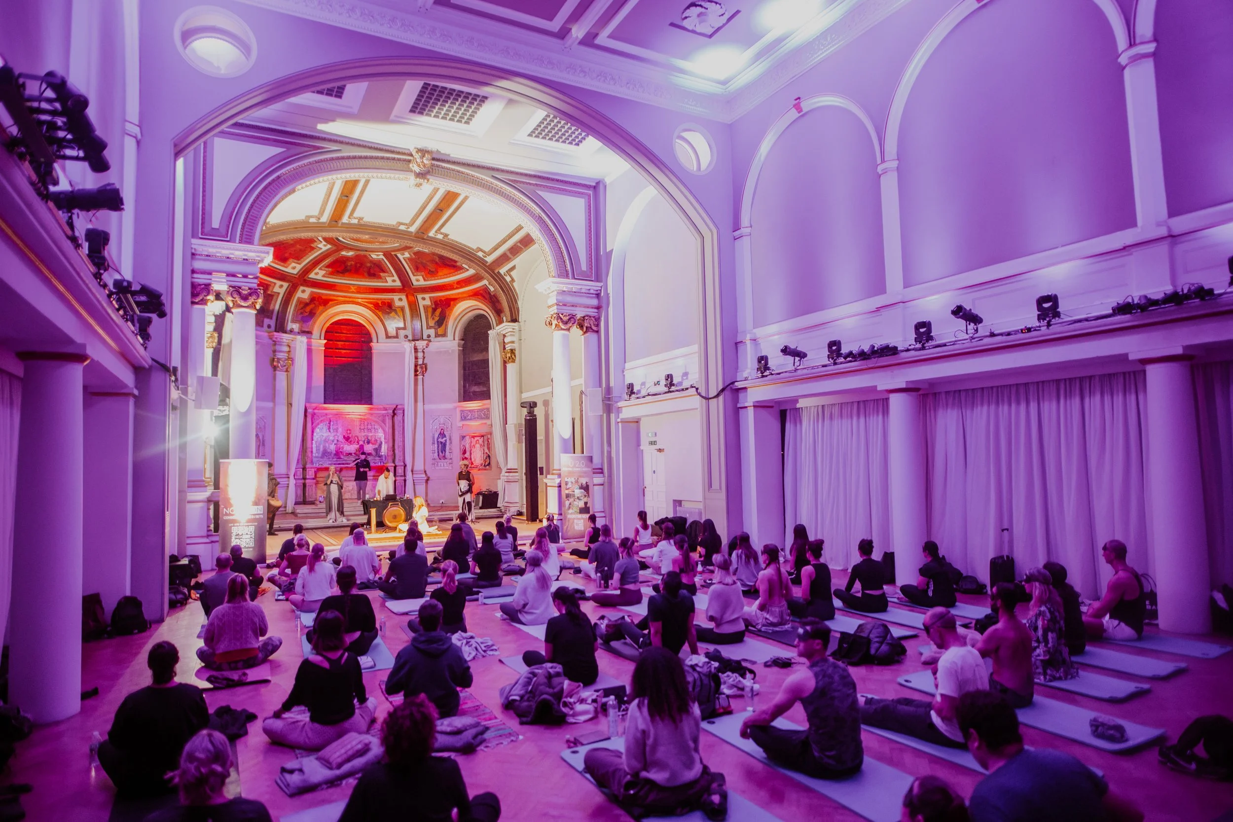 People participating in a meditation session inside a grand, ornately decorated hall with purple lighting.