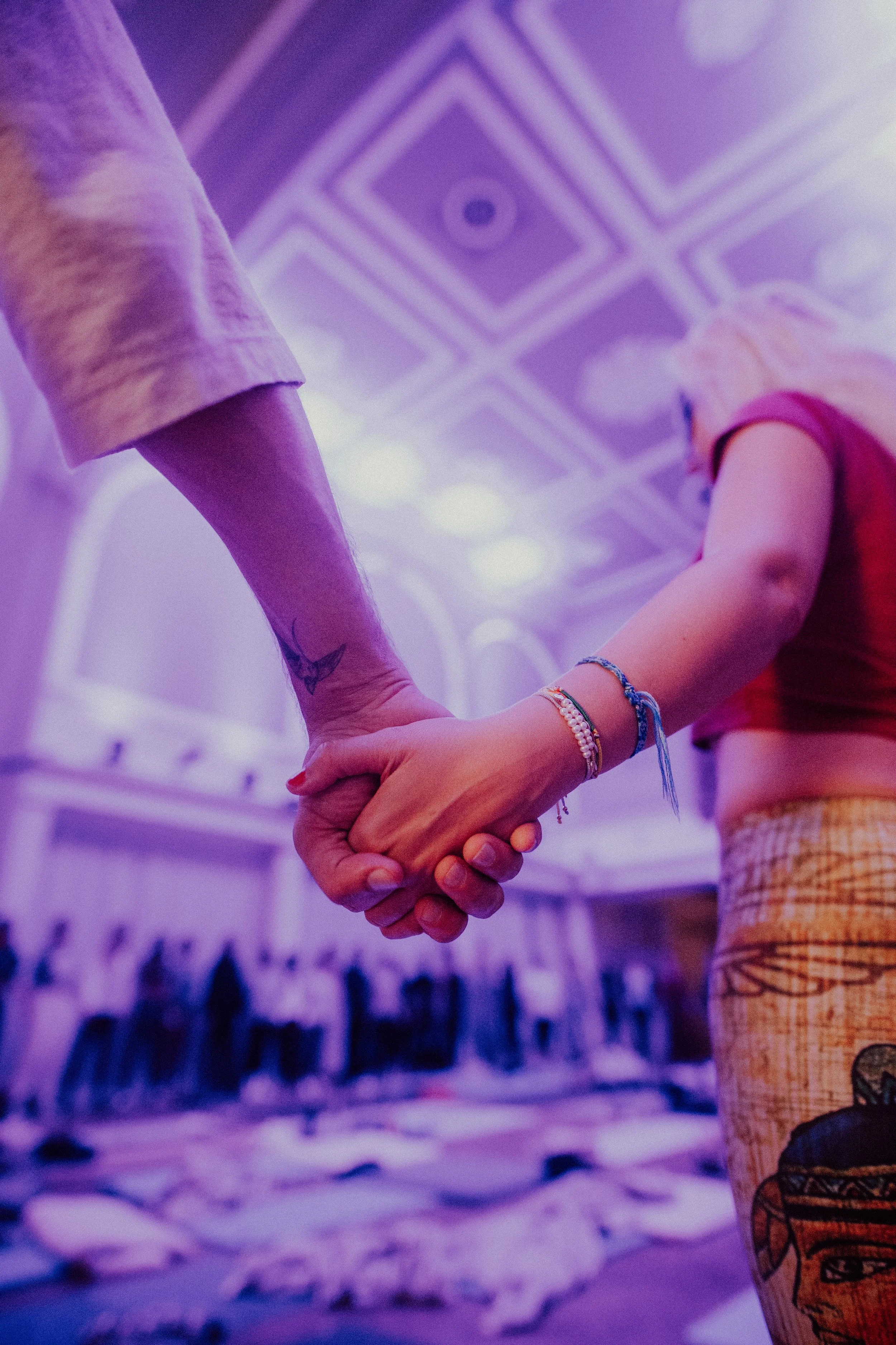 A close-up of two people holding hands, with a blurred background of an interior space with high ceilings and chandeliers, illuminated by purple lighting.