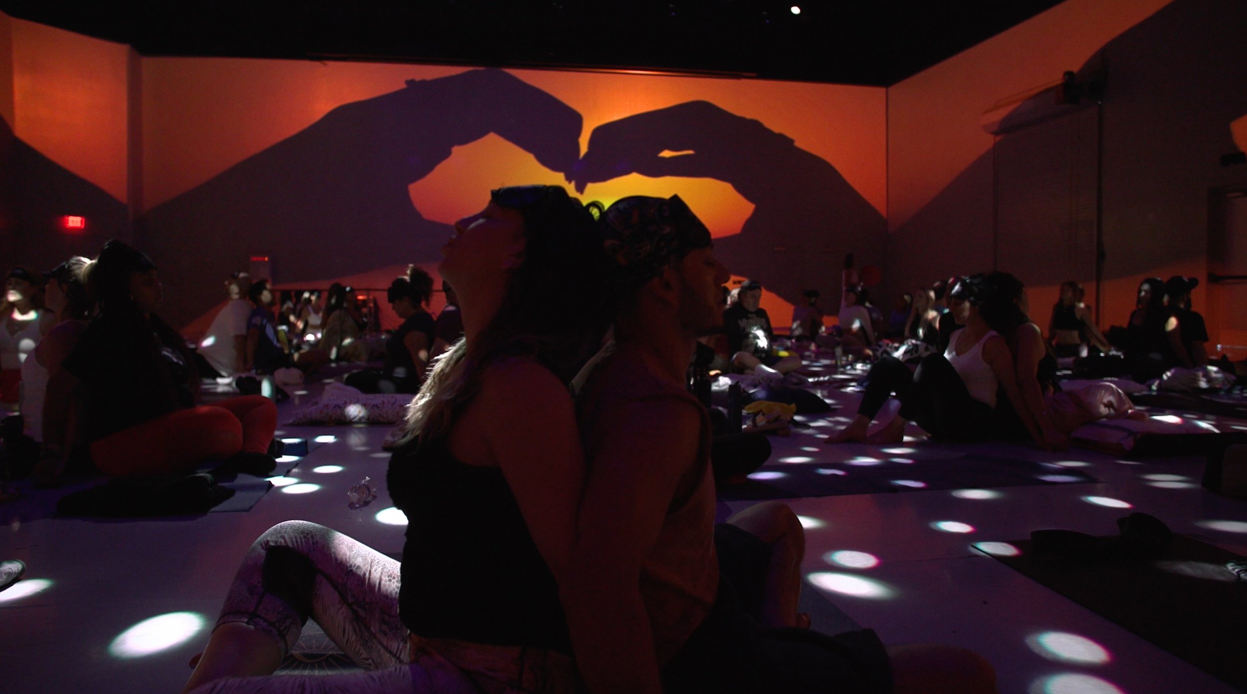 People participating in a yoga class in a dark room with a projection of a sunset and hands forming a heart shape on the wall.