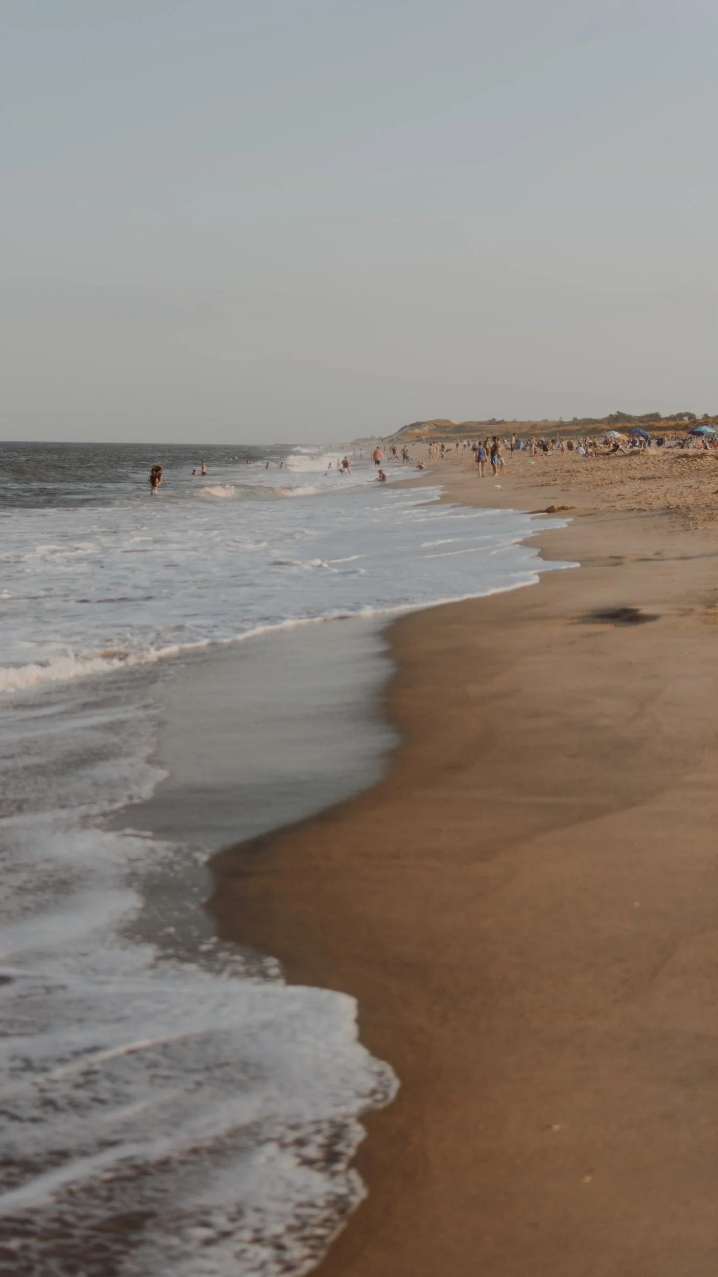 Beach scene with people swimming and relaxing on the sandy shore, waves crashing, and distant cliffs under a clear sky.