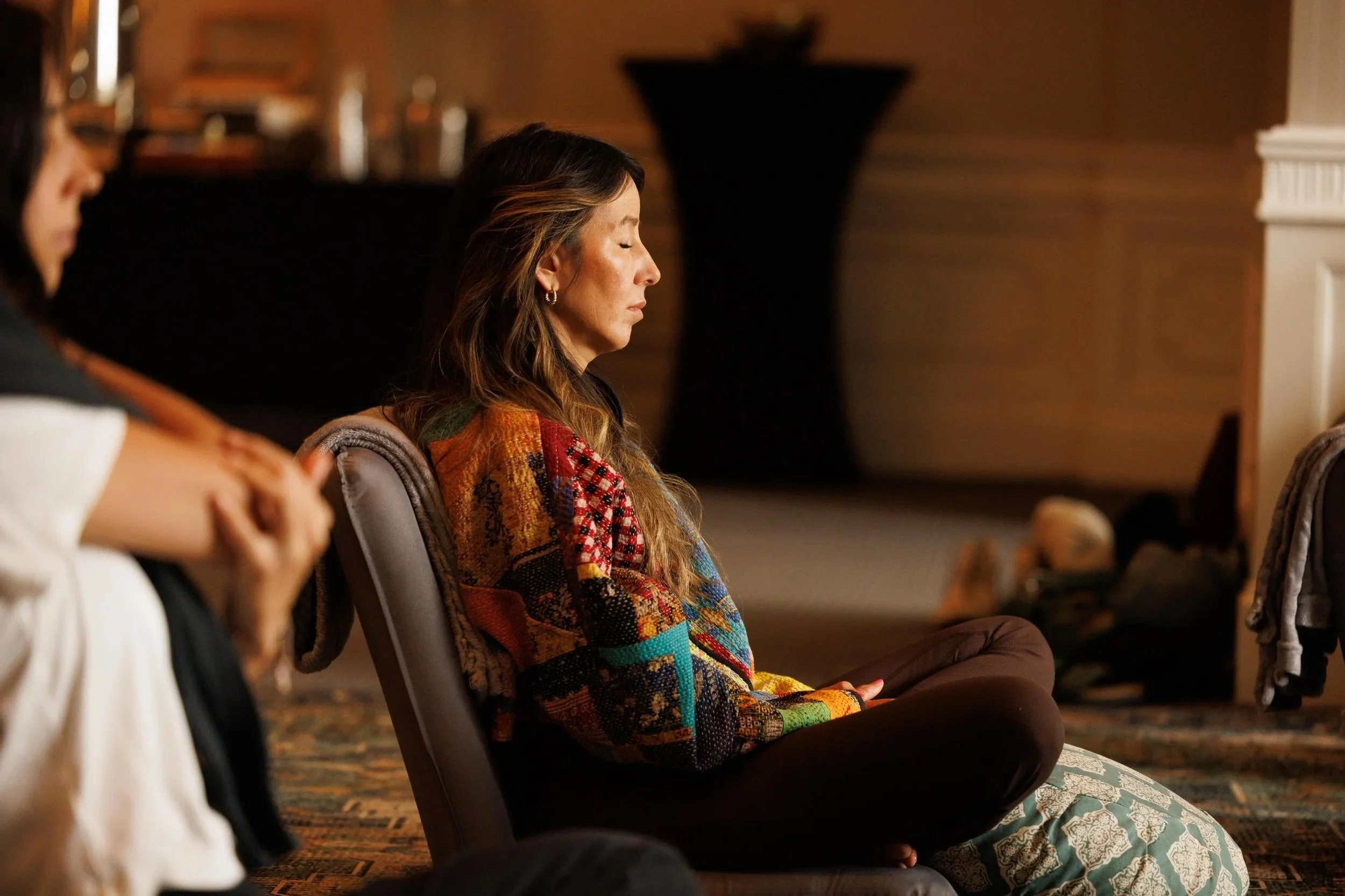 A woman with long hair, eyes closed, sitting cross-legged and meditating in a dimly lit room. She is wearing a colorful patterned sweater and patterned pants, with a calm expression.